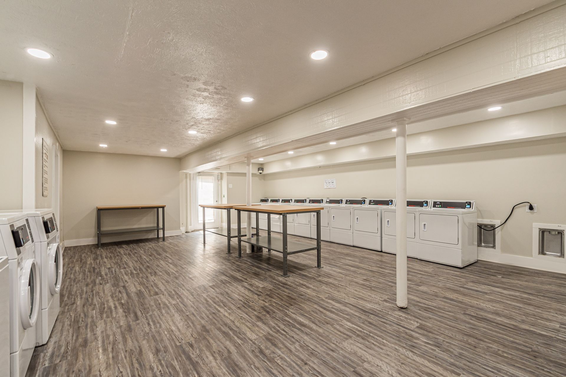 Empty laundry room with rows of washers and folding tables on wood-look flooring