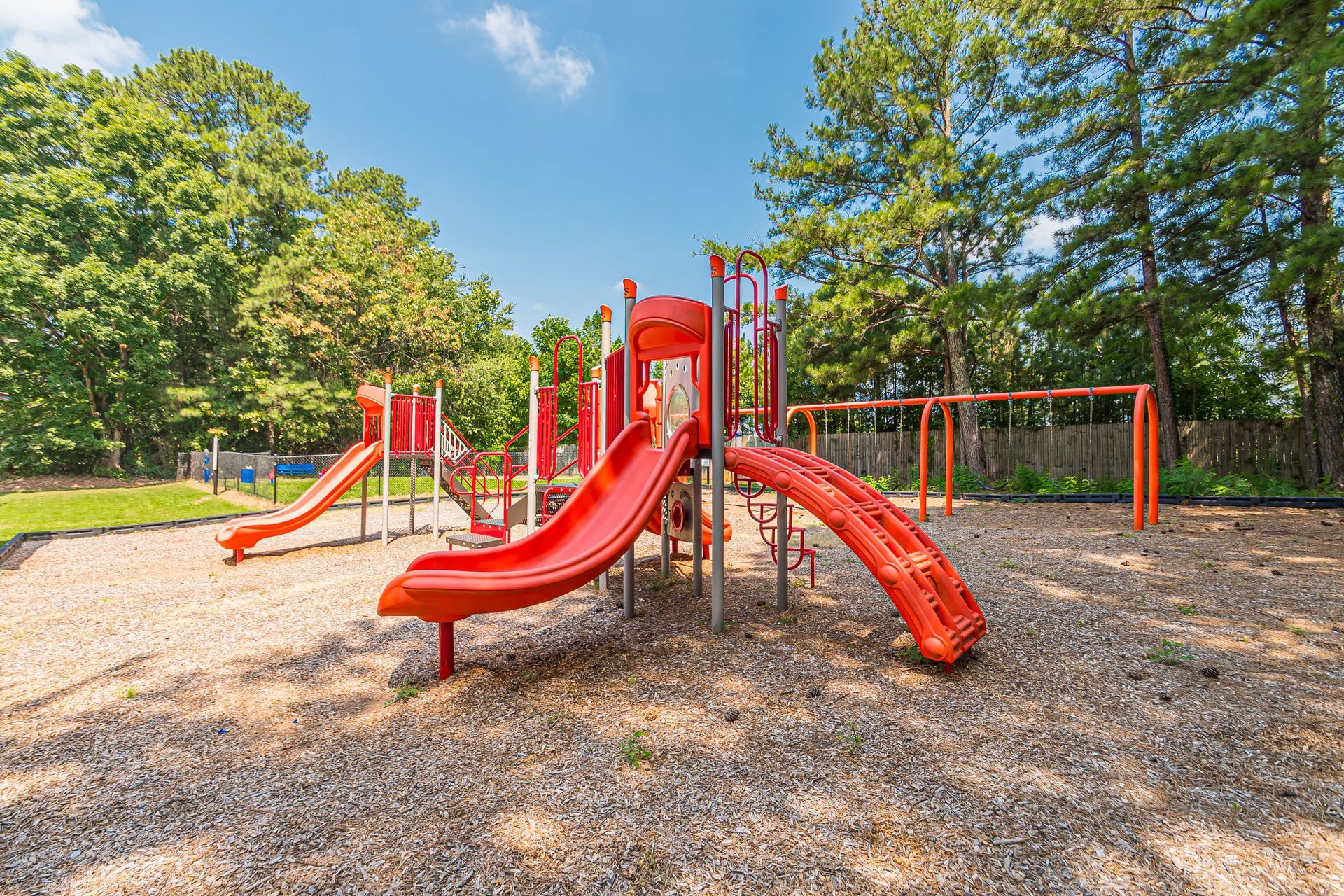 Red playground with slides and climbing structures on a woodchip park surface under a sunny sky.