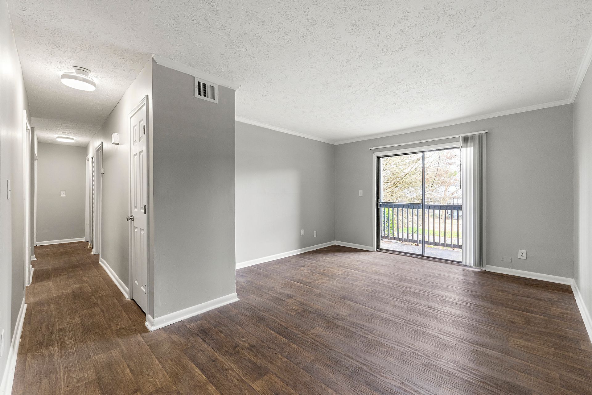 Empty apartment living room with gray walls, wood floors, hallway, and sliding glass door to balcony.
