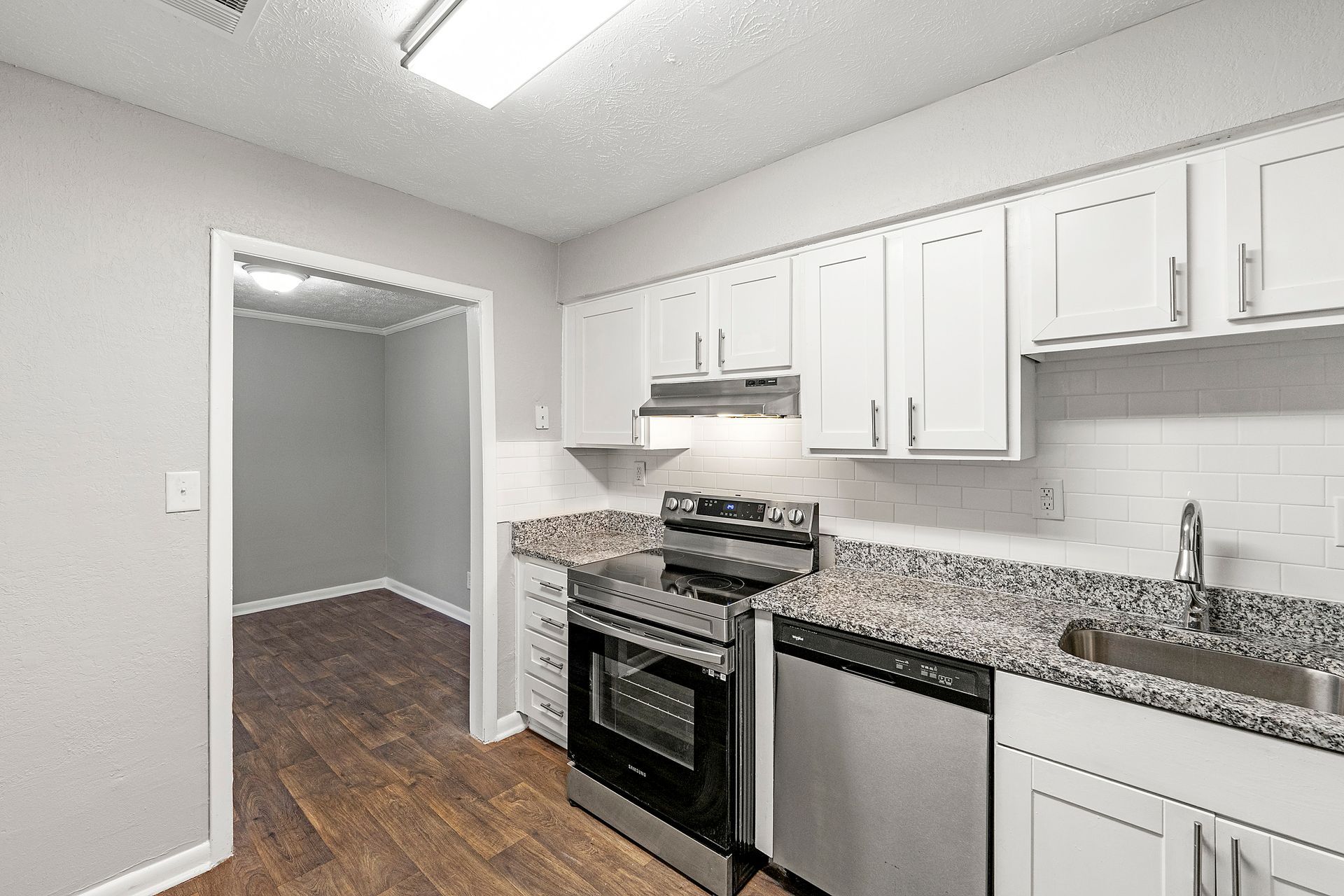 Bright white kitchen with granite counters, stainless appliances, and wood flooring leading to an open doorway.