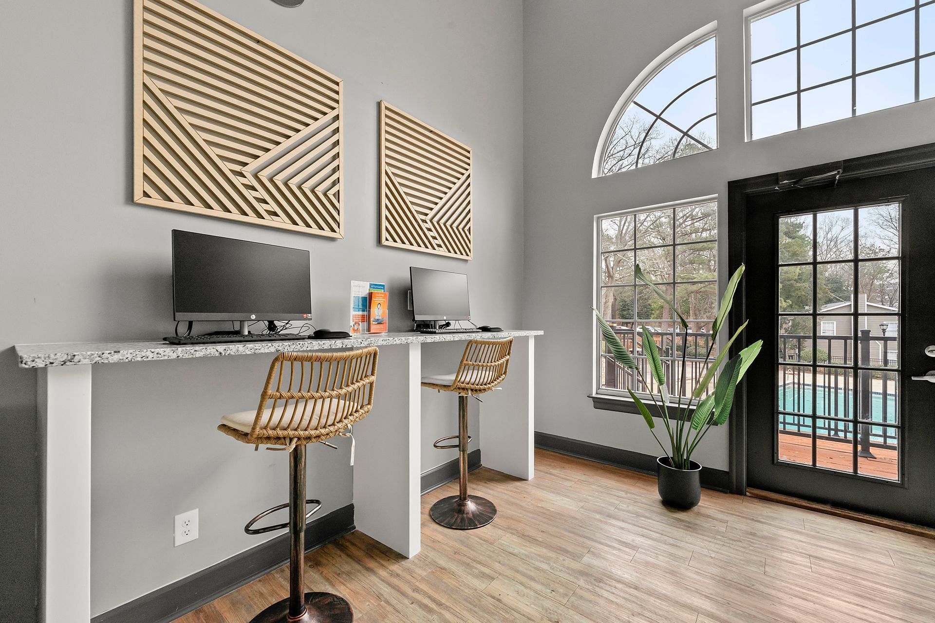 Modern home office with two desks, computers, woven stools, and large arched windows by a glass door.