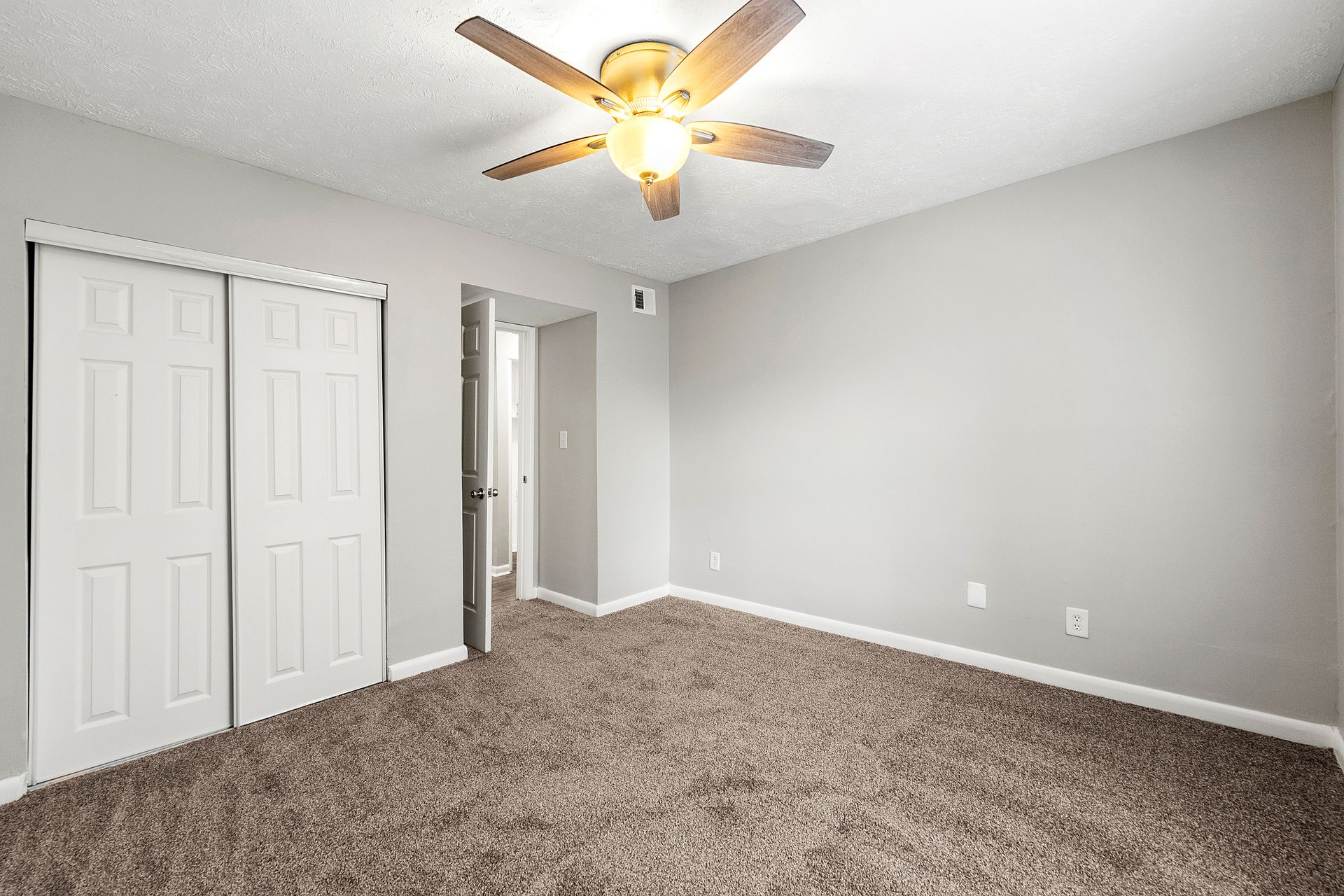 Empty carpeted bedroom with gray walls, white closet doors, and a ceiling fan on.