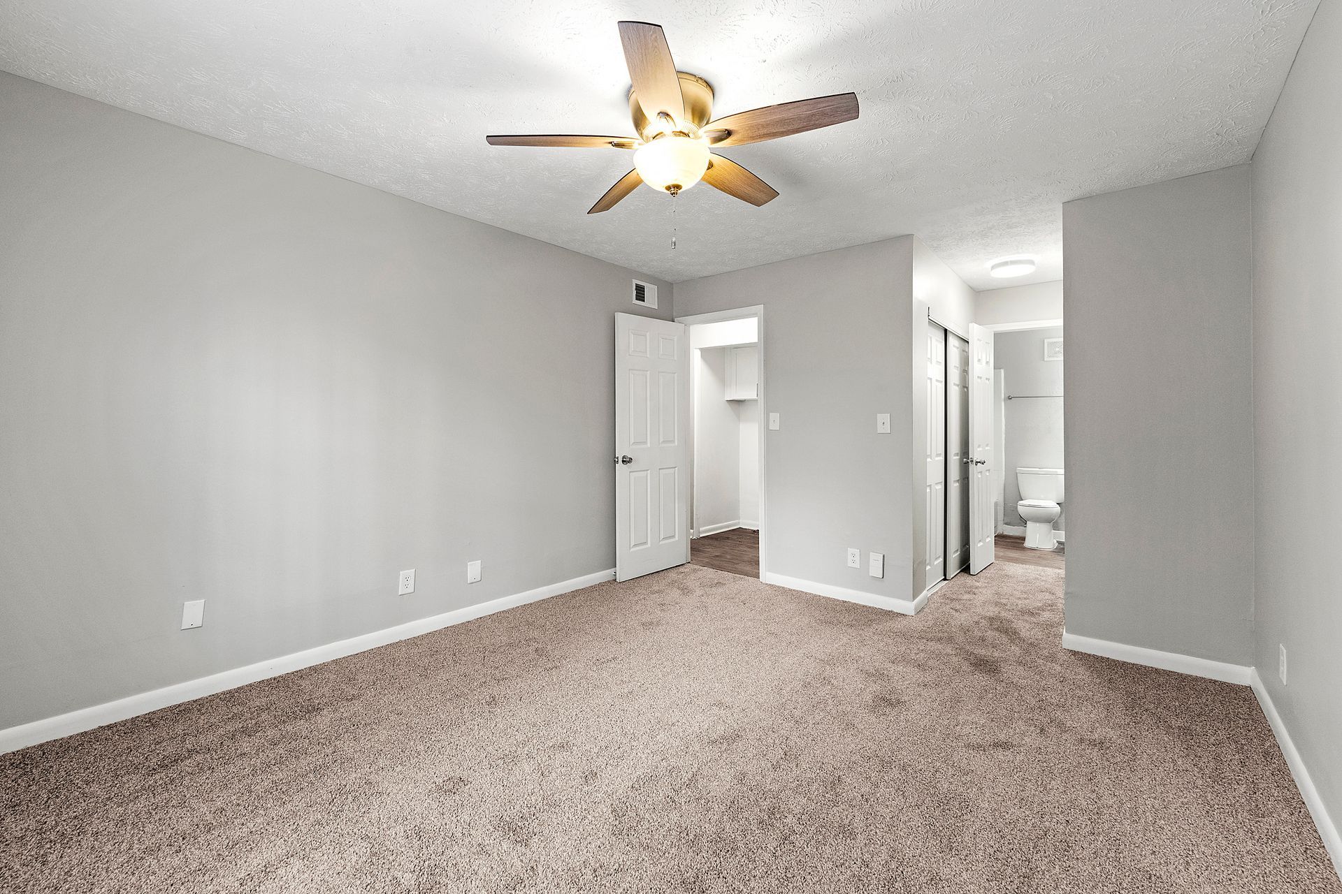Empty gray carpeted room with ceiling fan, open doorways, and hall view