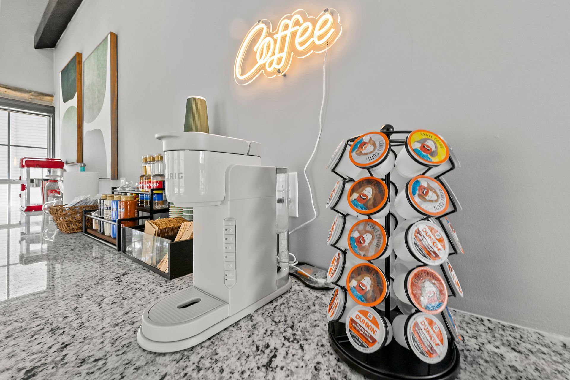 Coffee station with white machine, neon “Coffee” sign, and pods on a granite countertop