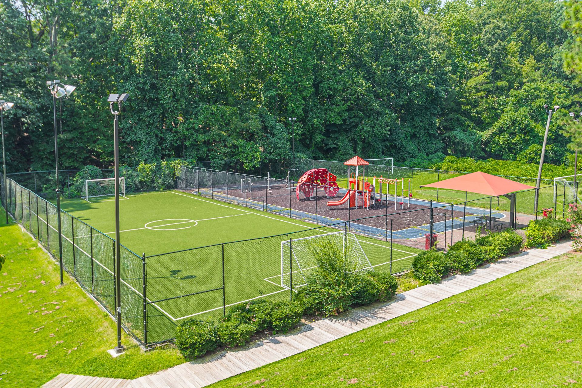 Fenced outdoor playground with soccer field, play equipment, and orange shade canopies in a green park