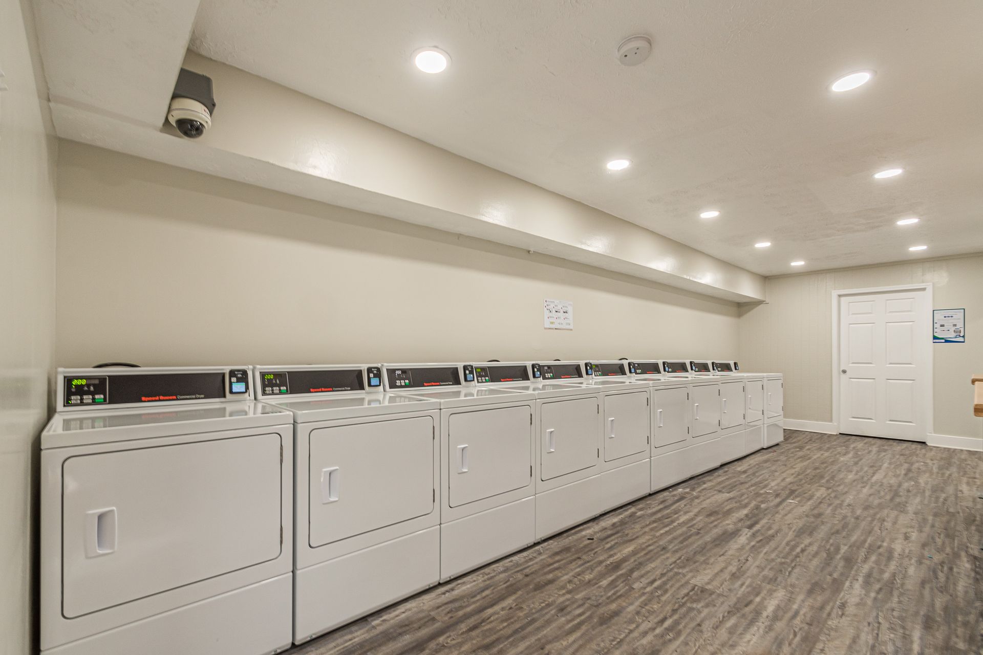 Laundry room with white washers and dryers lined along a wall, wood-patterned floor, and recessed ceiling lights