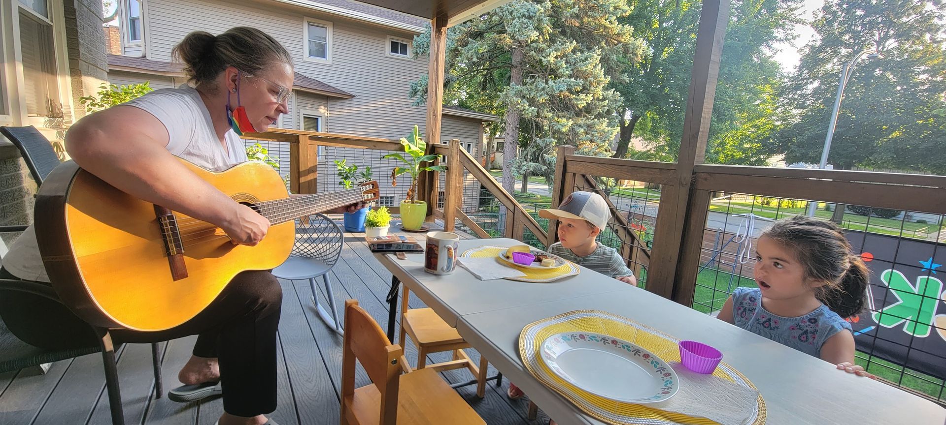 A teacher is seated on an outdoor porch playing a guitar while a young child sits at a table in front of her, eating food at a place setting.