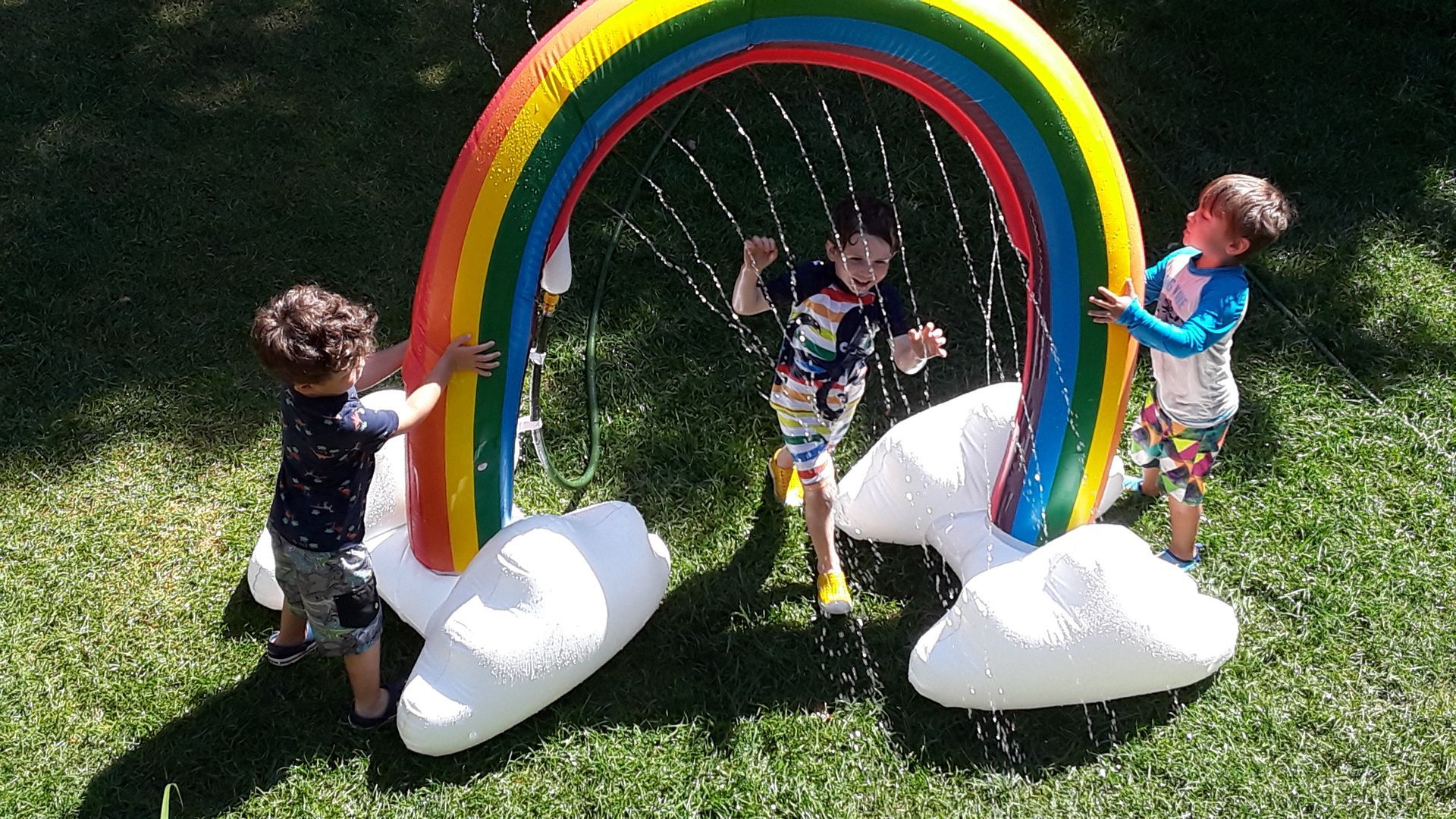 Two children hold each side of an inflatable rainbow that is also a backyard sprinkler toy. A third child is running under the rainbow, through the sprinling water.
