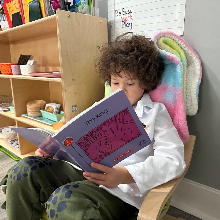 A young child seated comfortably by themself in a playschool setting, looking at a large open storybook.