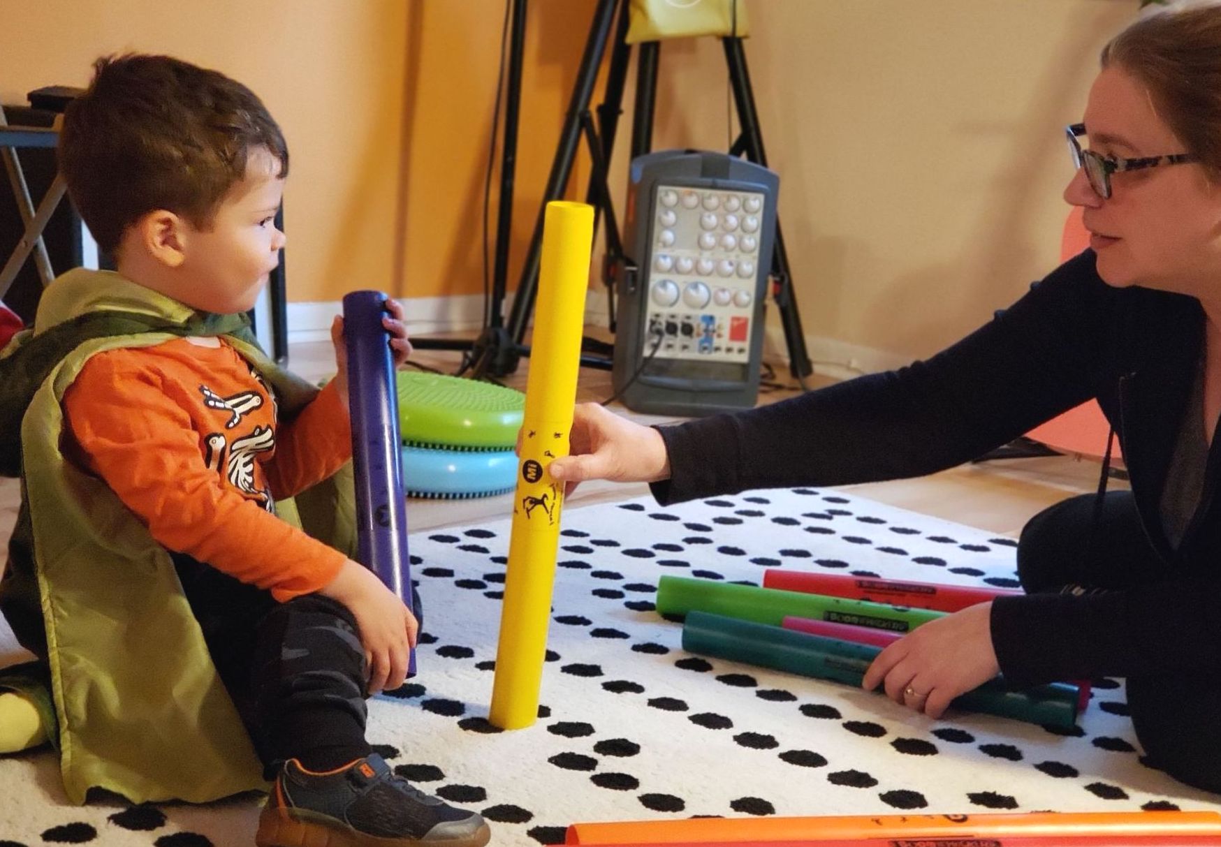 An adult teacher sits on the floor accross from a young child. The teacher is holding a piece of yellow tubing, while the child is holding a piece of blue tubing. There are other tubes of different colors on the floor.