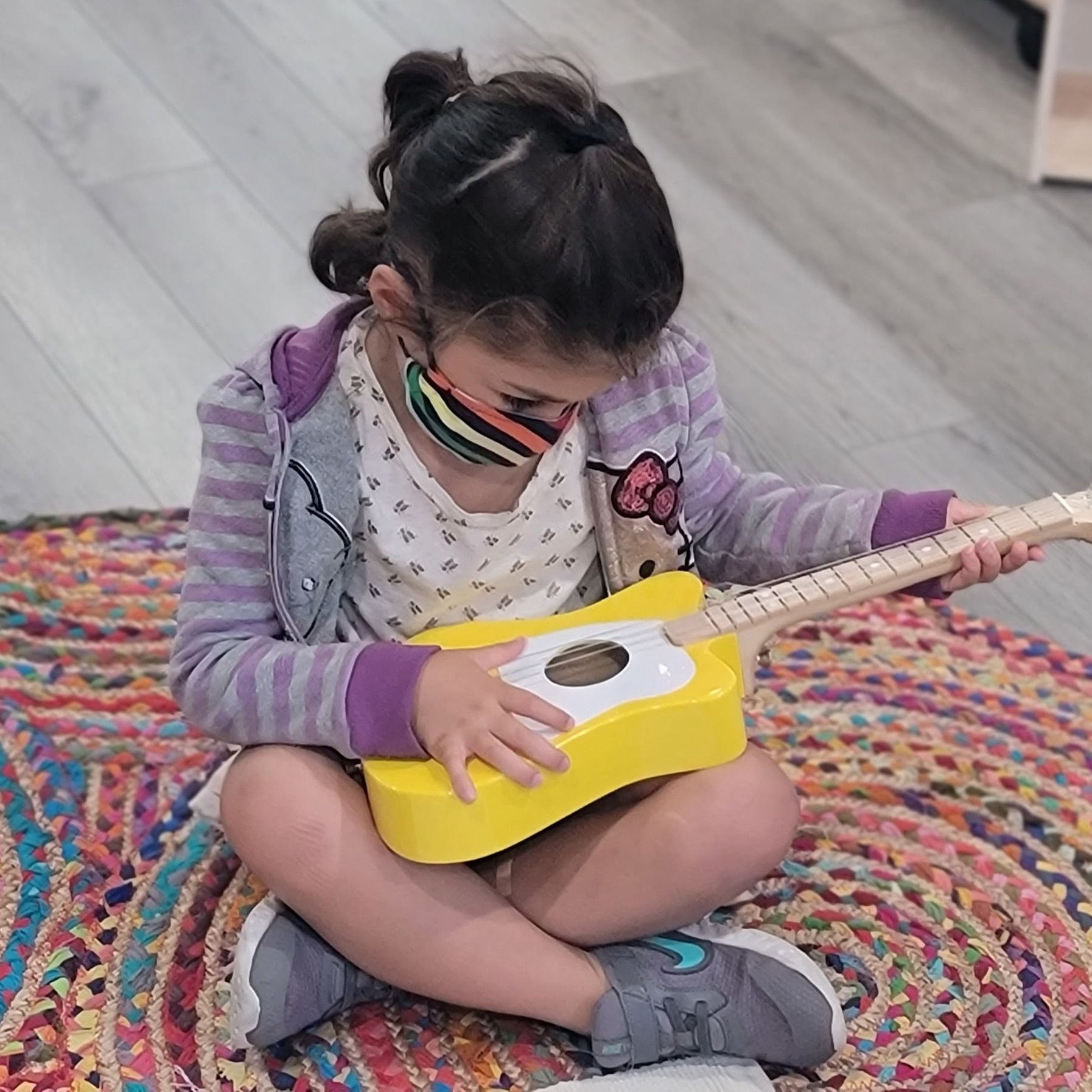 A child examines a small yellow child-sized guitar while seated on a colorful rug.
