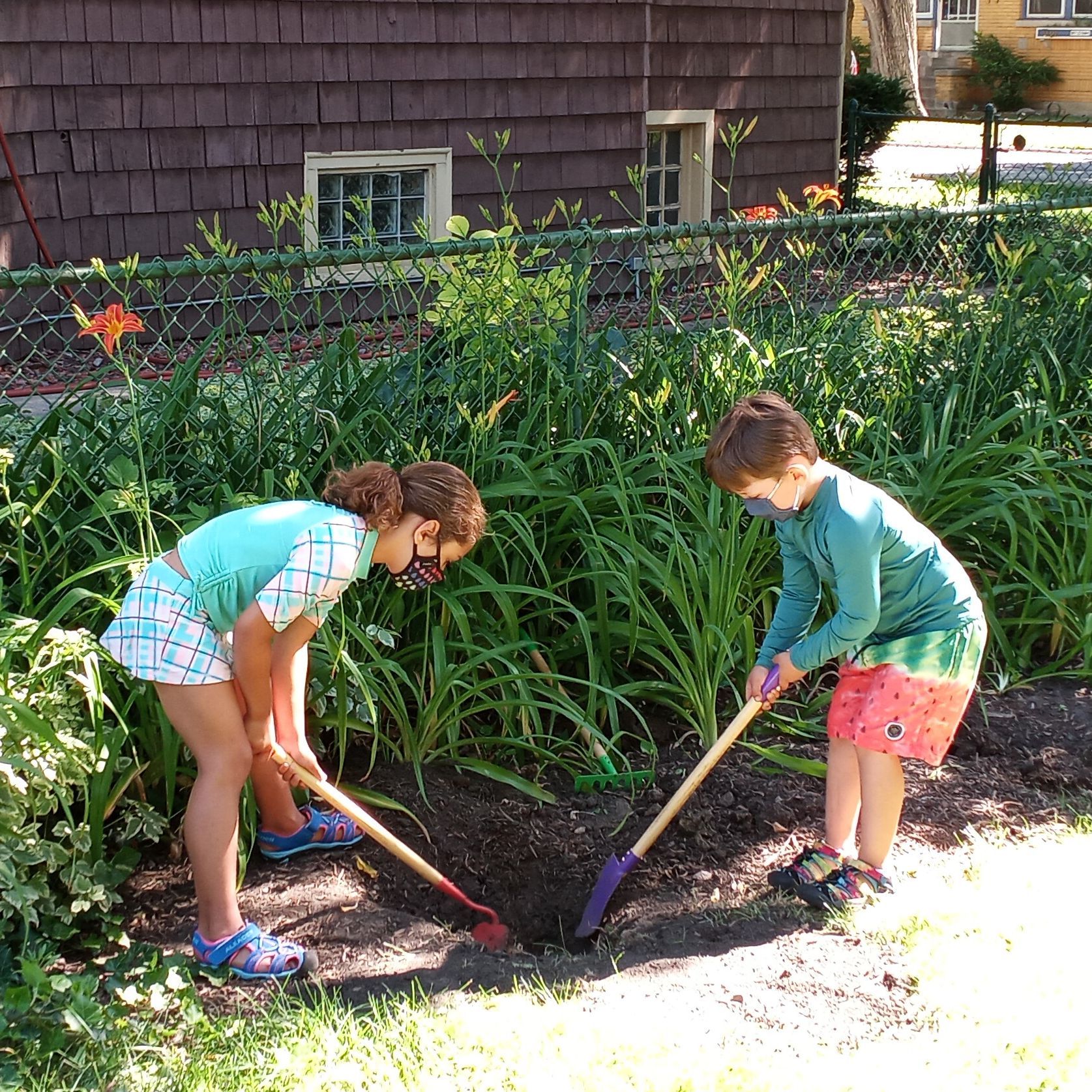 Two children are outdoors bending over to dig in a dirt patch with long handled garden tools.