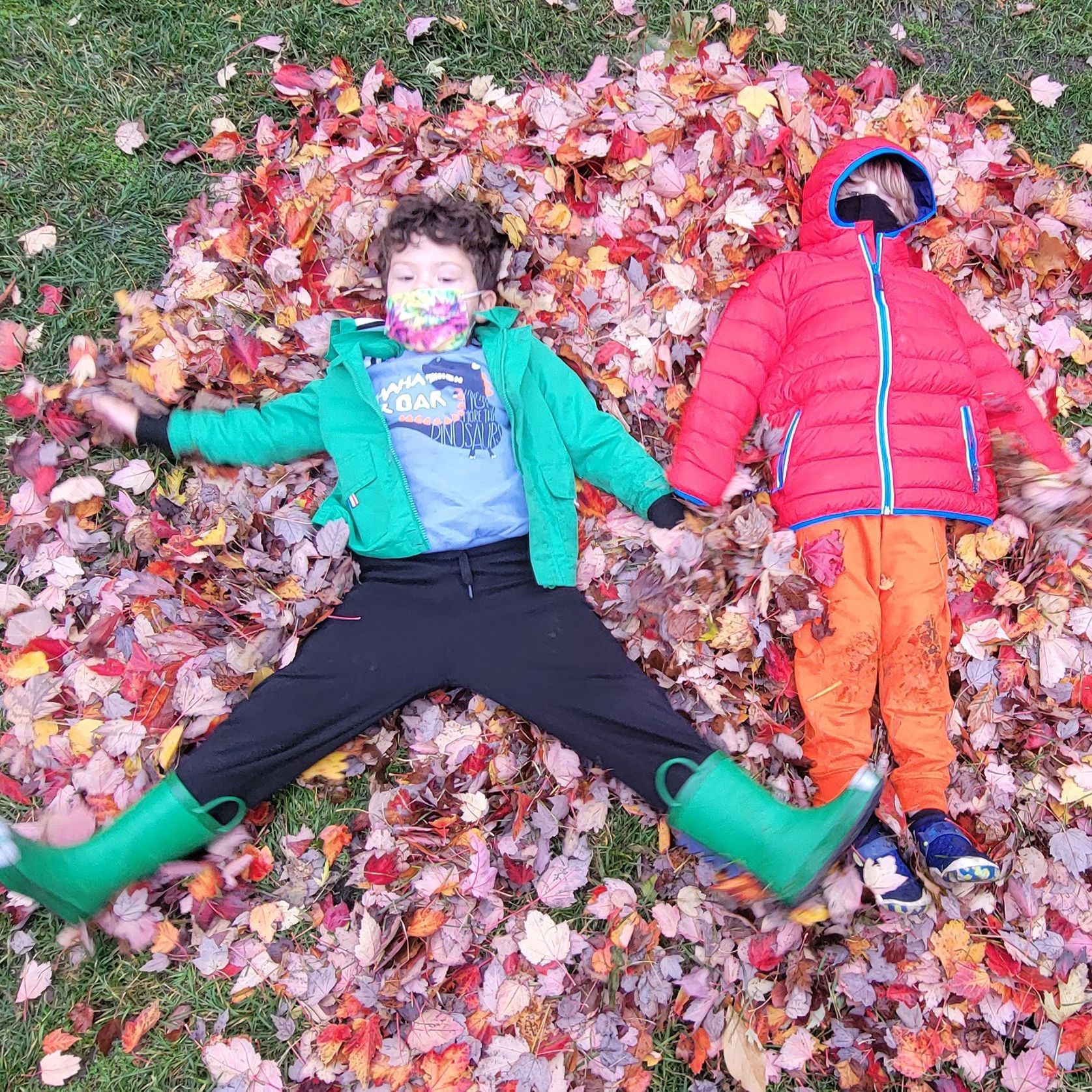 Two children are lying outdoors on their backs in a pile of autumn leaves. They are wearing jackets.