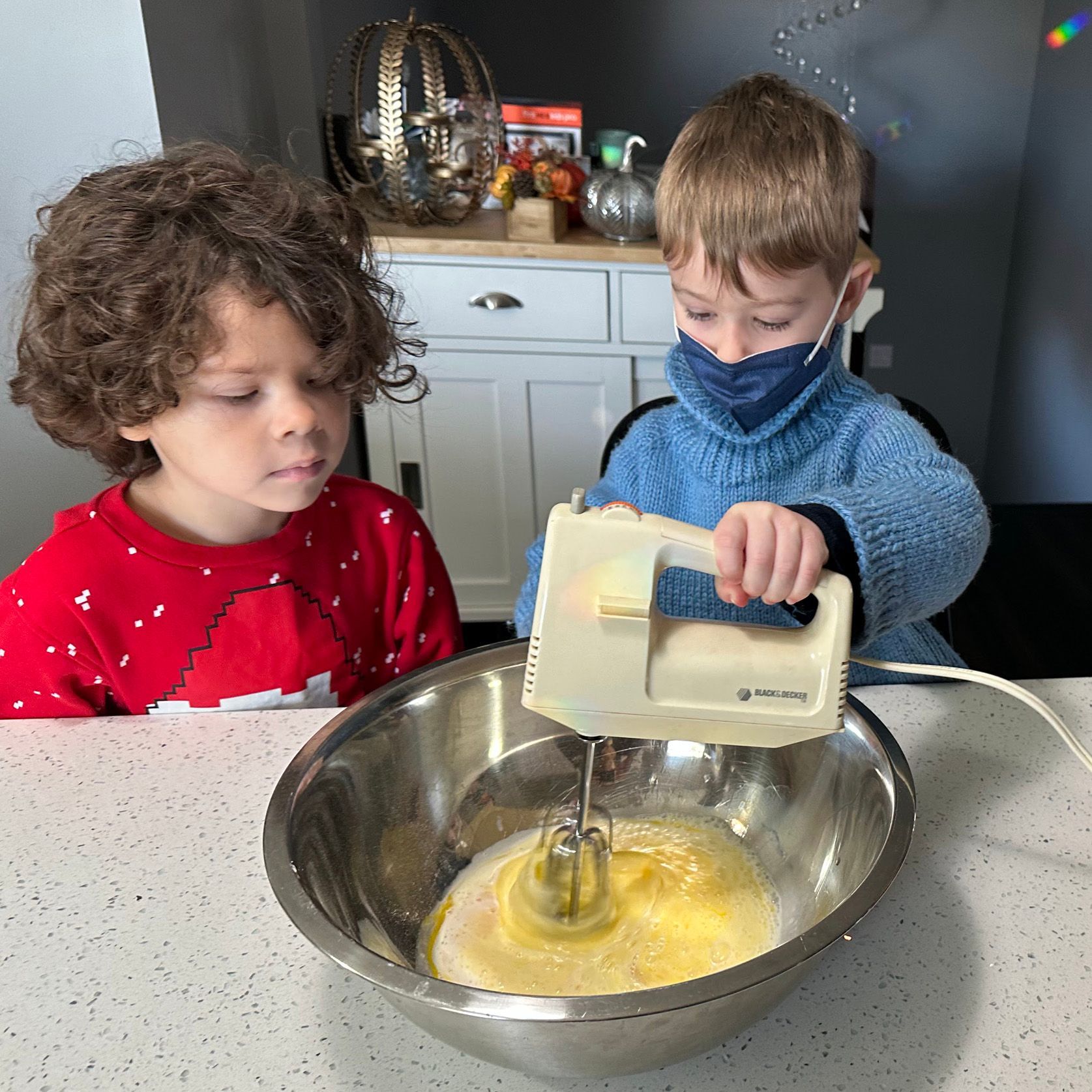In a kitchen, two children are standing at a table. One child looks on as the other child uses an electric mixer to beat eggs in a steel bowl.