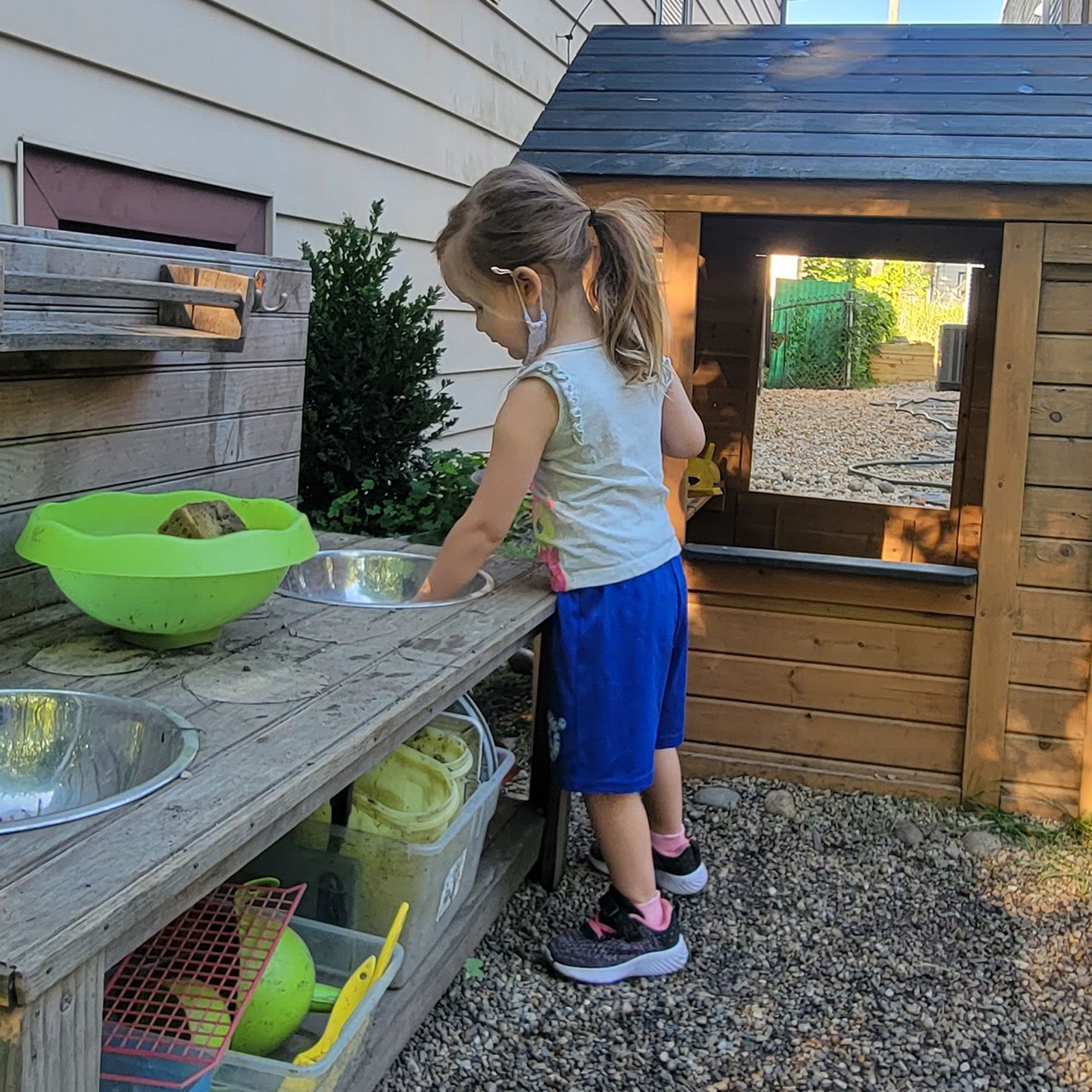 A child stands at an outdoor table that is equipped with steel basins and plastic bowls. She is busy with one of the basins.