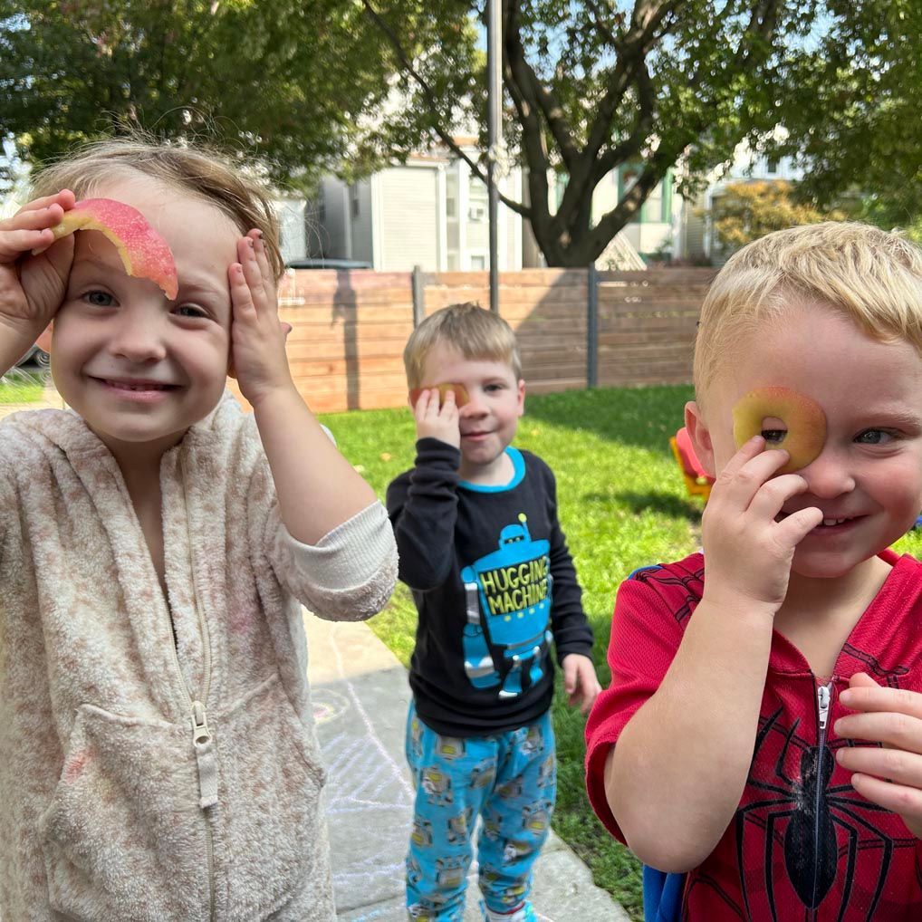 Three children smiling. Each child is holding an orange leaf over one of their eyes.