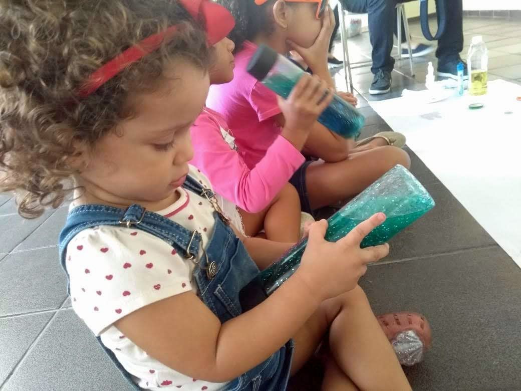 Three small children sit in a row on the floor holding lidded jars of colorful liquid. They are playing with the jars to watch the colored liquid flow.