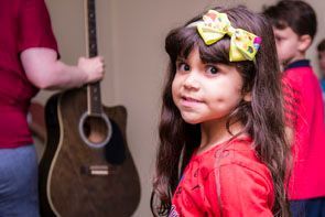 Child with bow in hair near an adult holding a guitar in a music class setting.