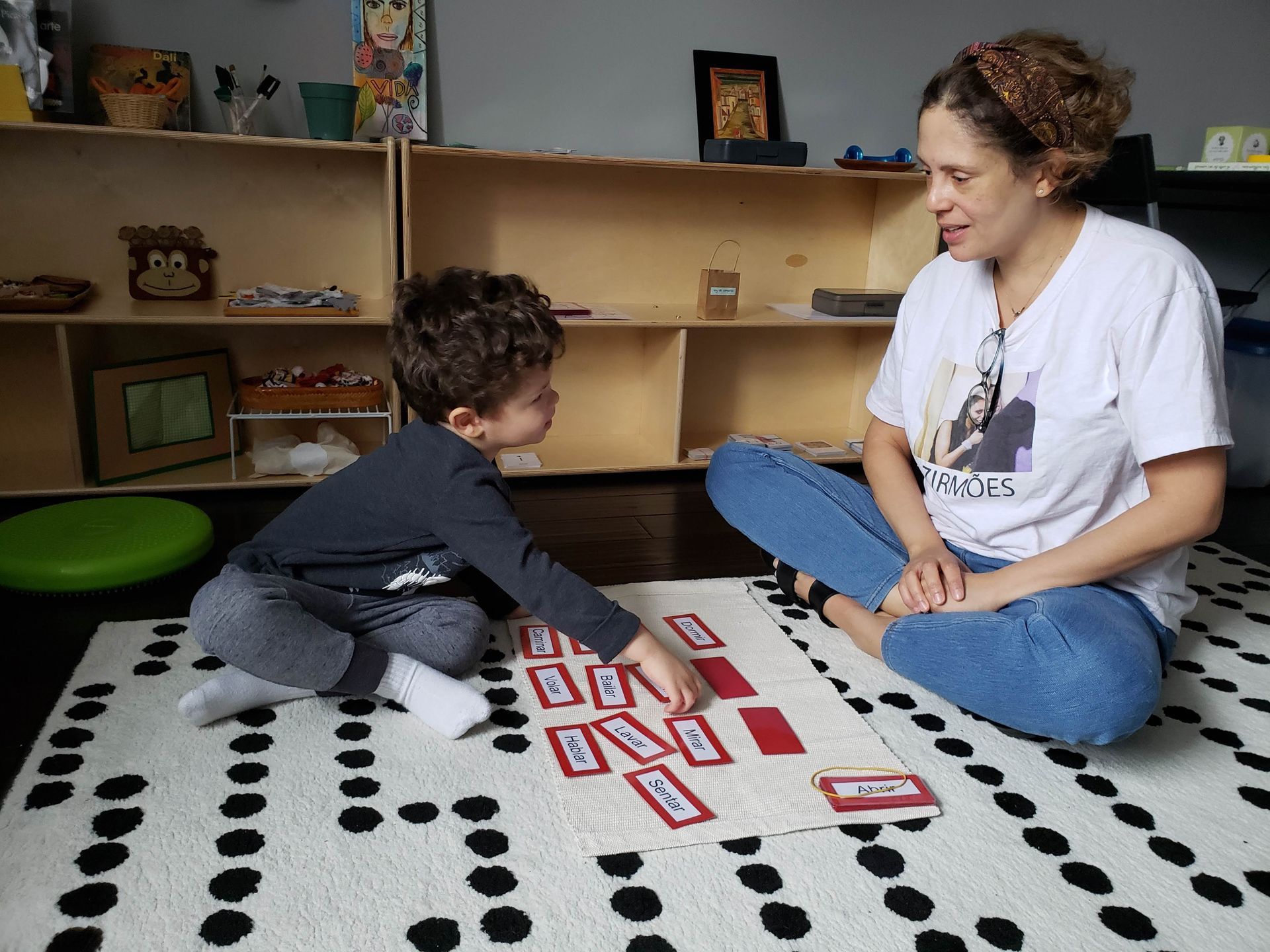 Teacher and young child sit across from each other on a rug during a learning activity. the child is pointing to one of several cards that have words on them.