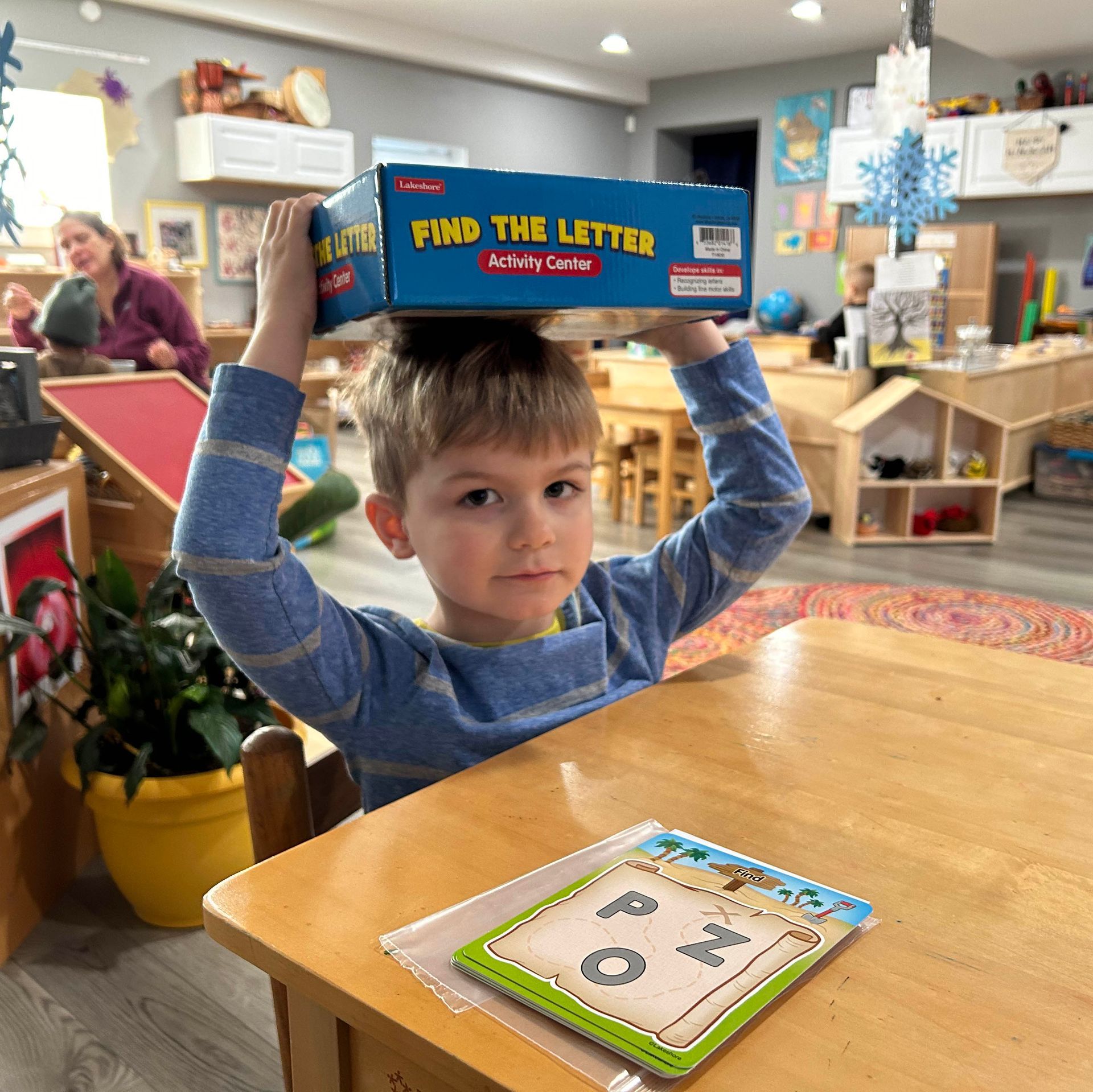 A young child seated at a table in a preschool setting holds a game box on top of his head.