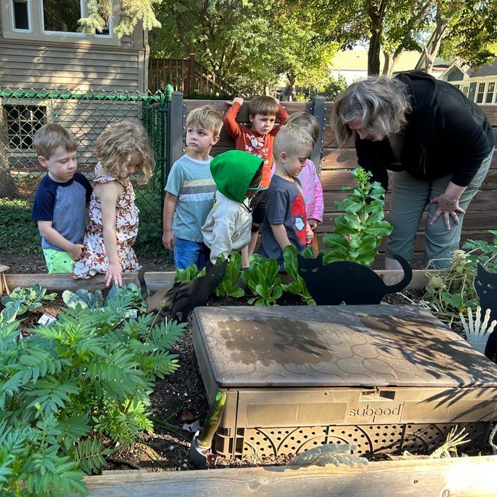 A group of children are with an adult women in a backyard setting. The children are in line next to a black cat as if to take turns petting the cat.