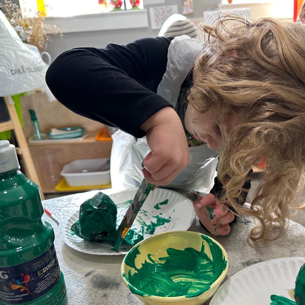 A child in an artsmock stands hunched over a small table, painting a small item with dark green paint. 