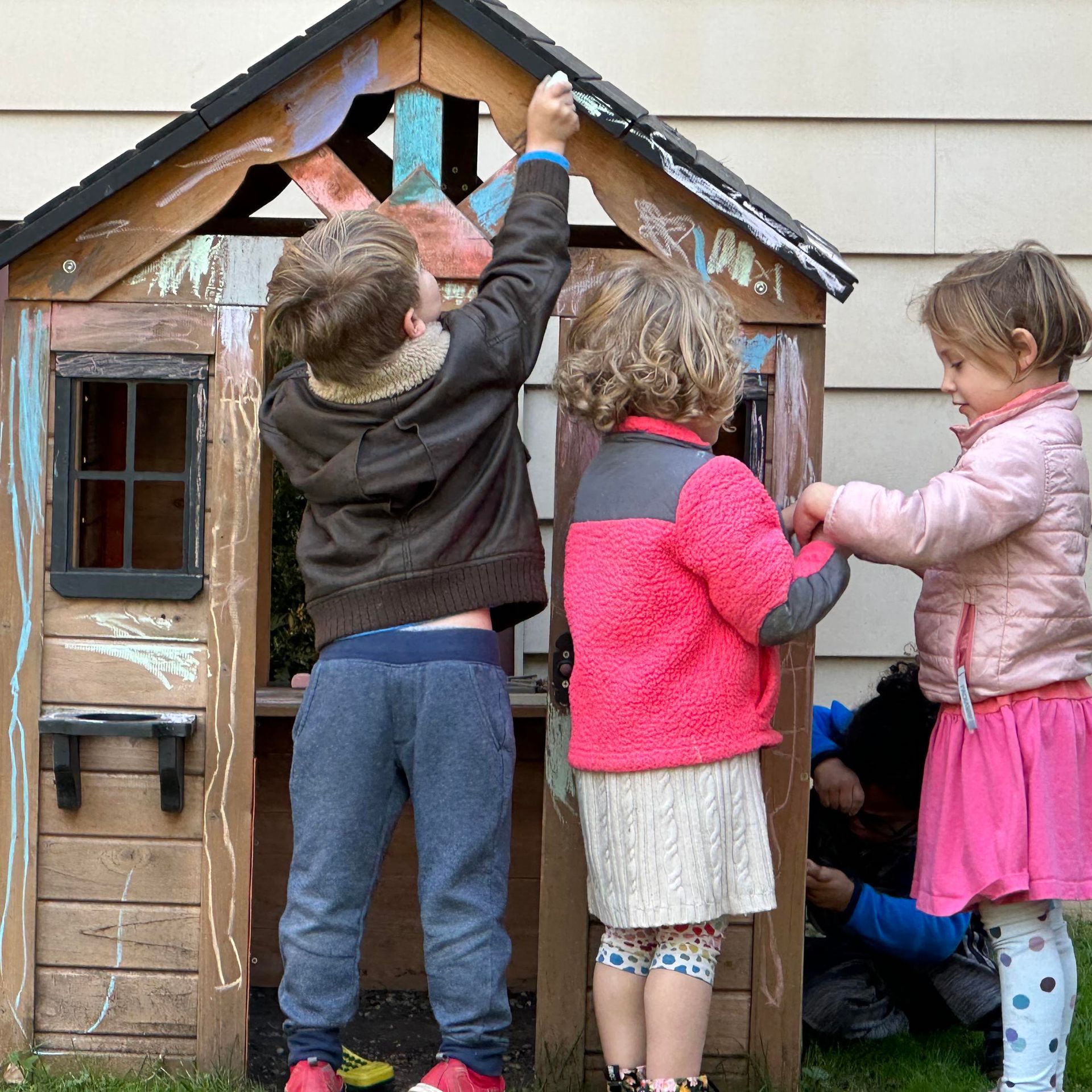 Three children are outdoors next to a wooden playhouse.