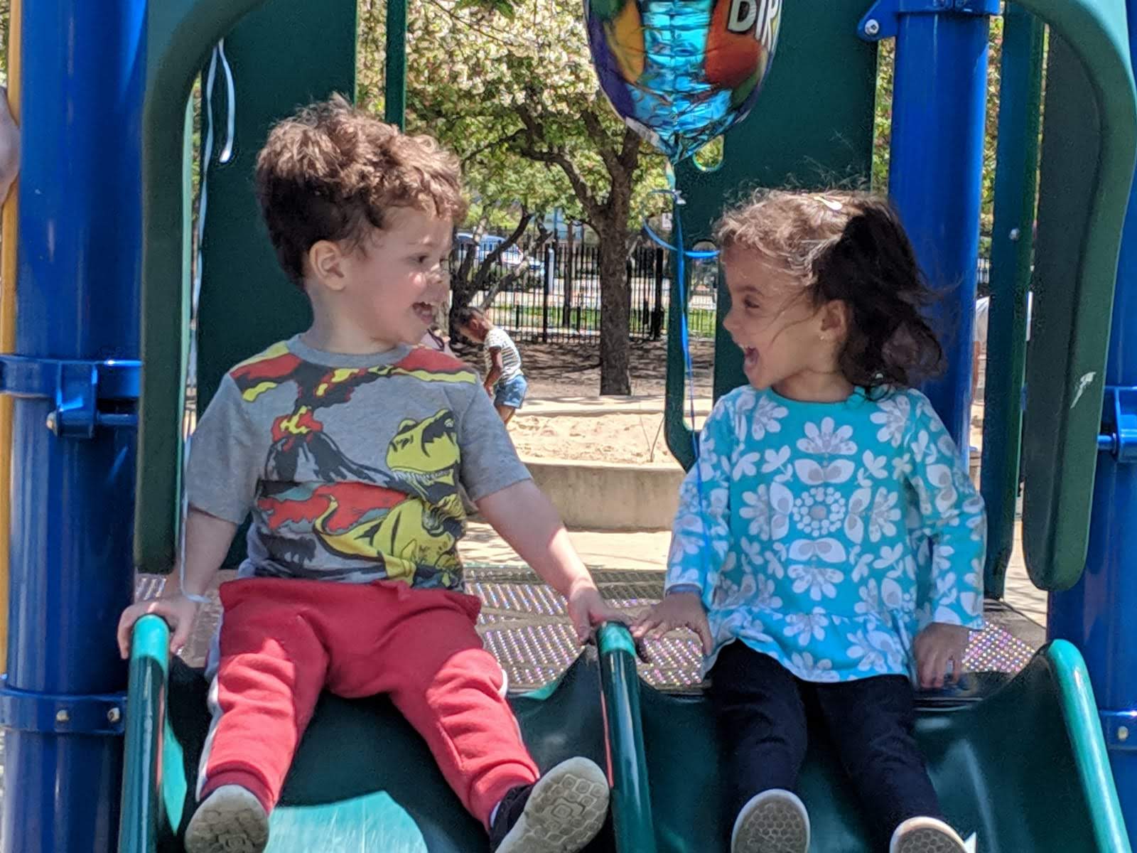 Two small children are at the top of a double slide. They are looking at each other, each prepared to go down their side of the the double slide.