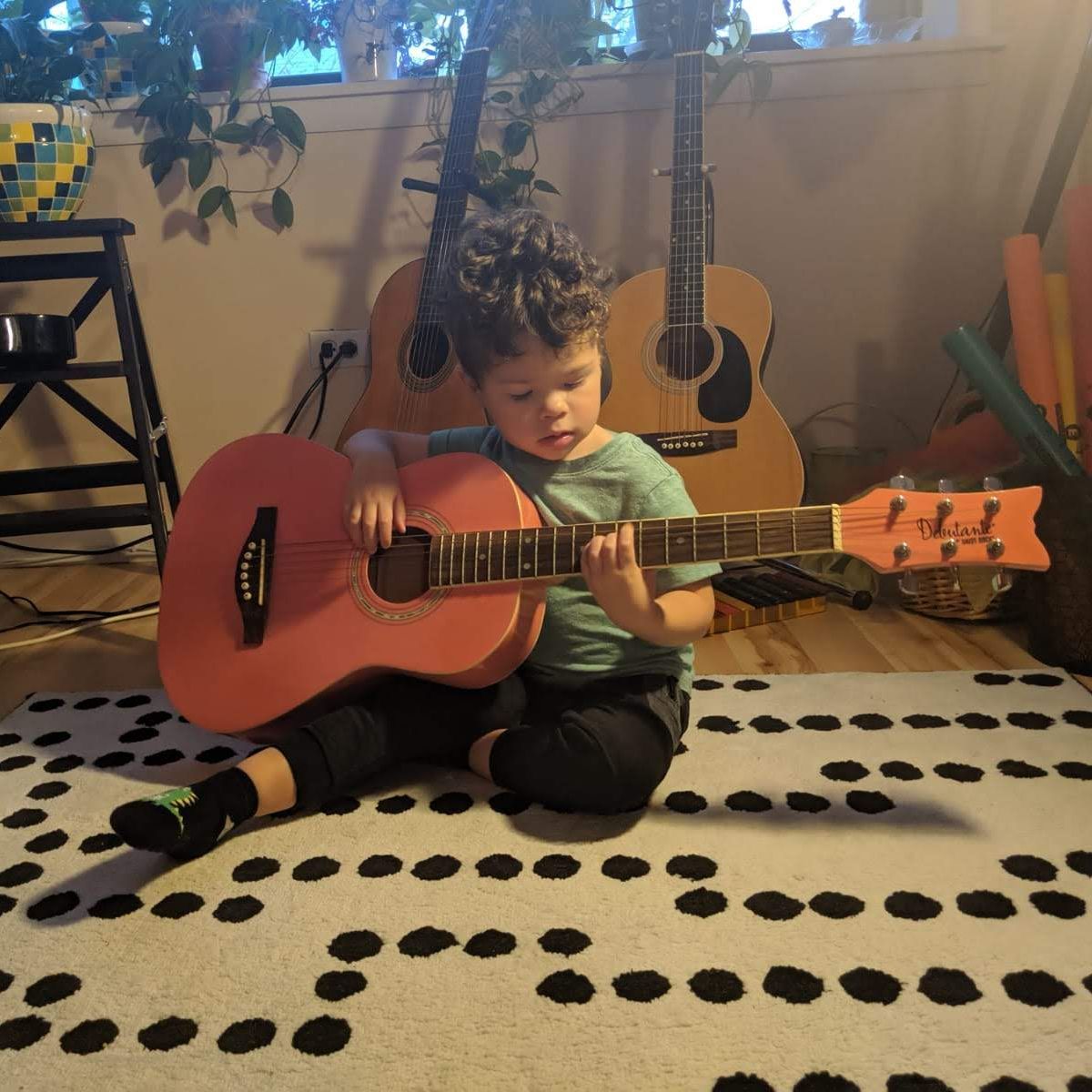 A young child sits on a rug on the floor holding and playing a guitar that is a bit large for them.