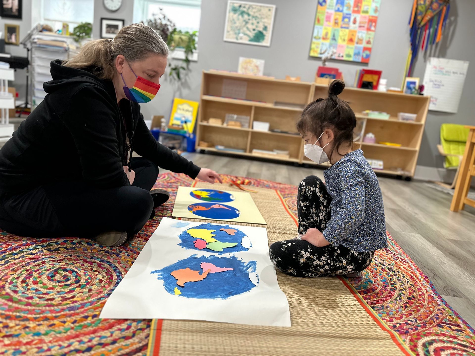 A teacher sits on the floor accross from a young child. There are painted pictures of the planet Earth between them that both are examining.