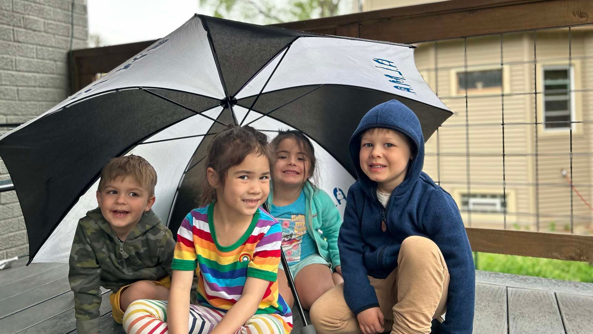 Four children are seated on a sidewalk under a large black and white golf umbrella.
