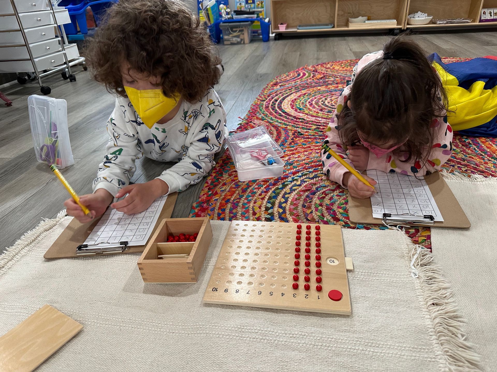 Two children with facemasks are in a preschool setting, laying on a rug while writing notes on a clipboard. in front of them is a board with an arrangement of marbles in three rows. The children appear to be engaged in a math activity with the marbles.