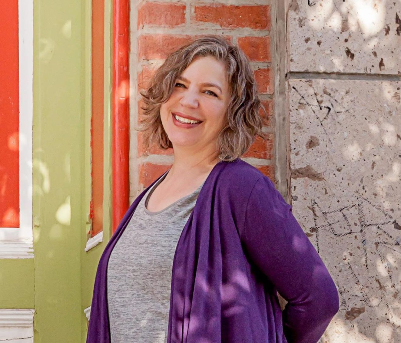 A smiling woman outdoors in a gray t-shrt and purple cardigan.