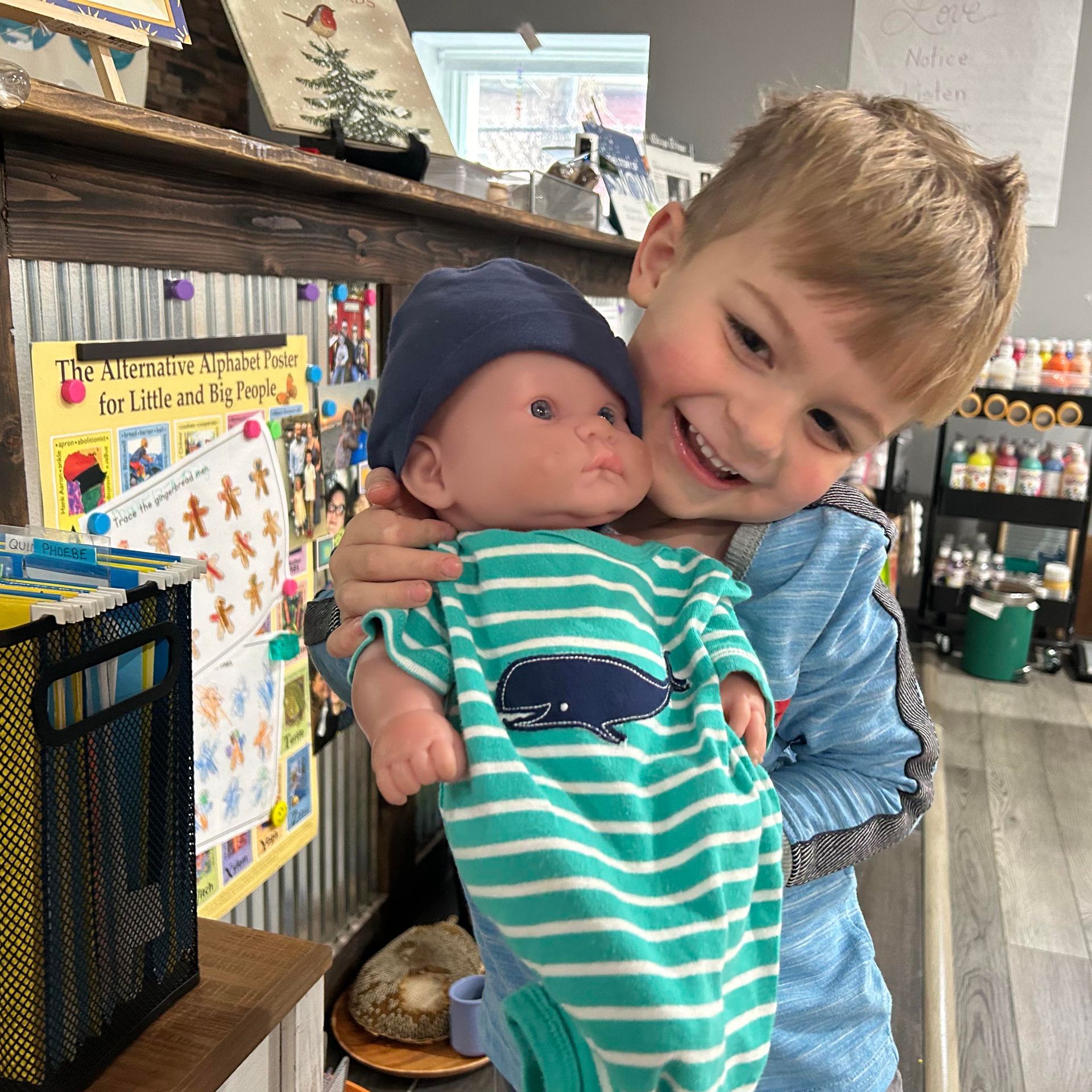 A smiling young child in a preschool setting holds a baby doll closely.