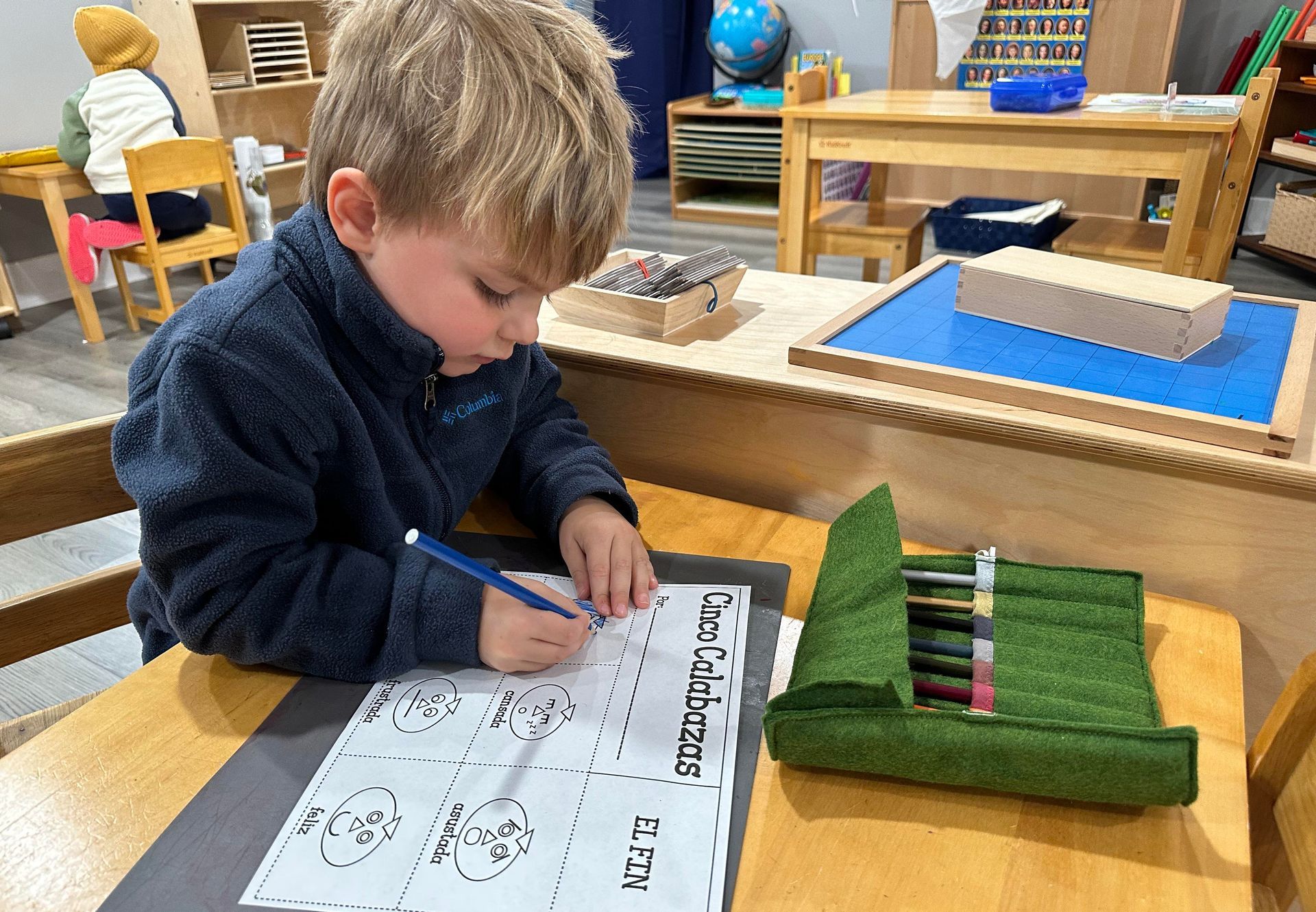 A young child seated at a low wooden table in a preschool enviornment is writing with a marker on a large worksheet.