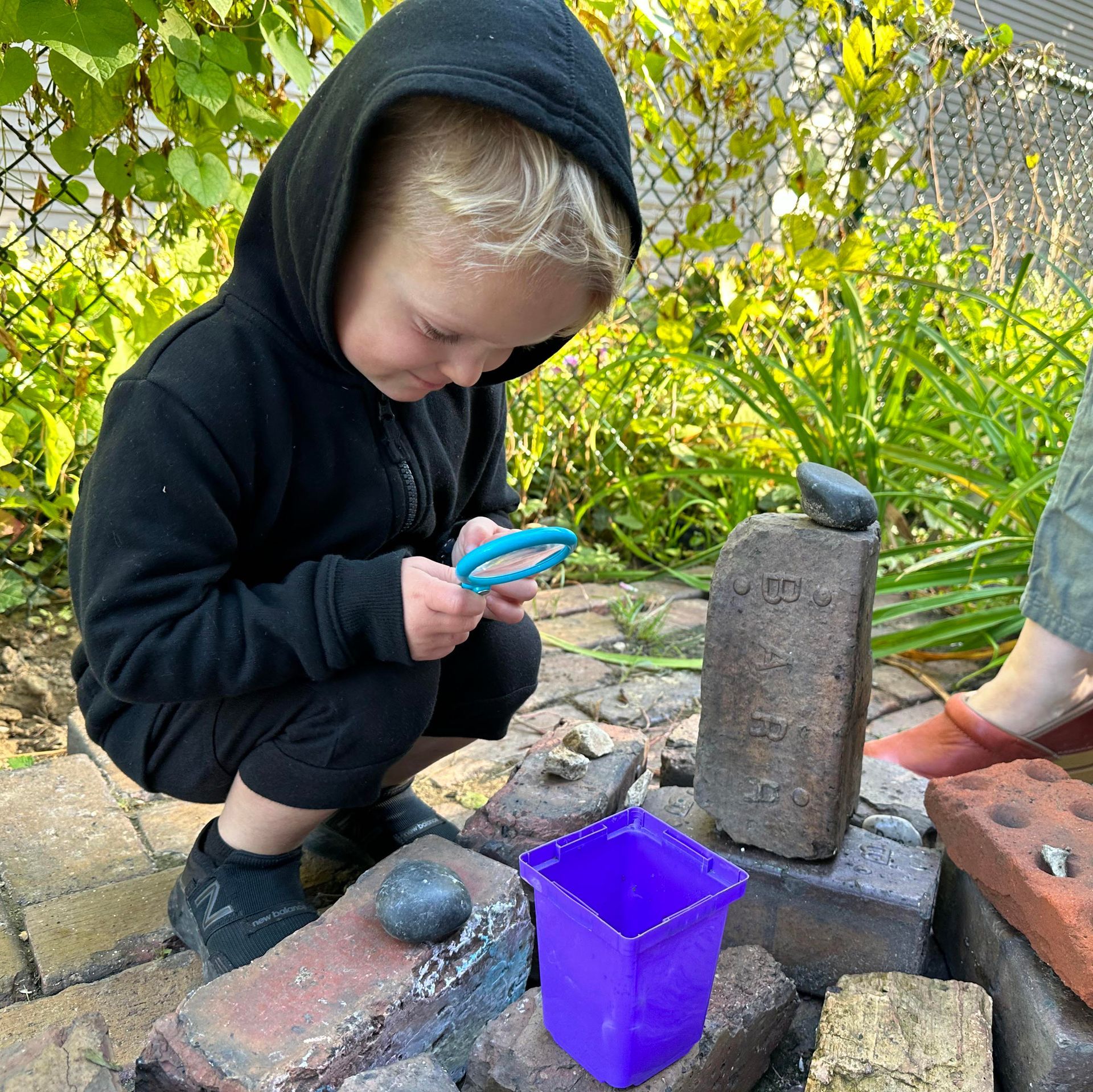 A young child wearing a hoodie is outdoors and squats to examine a group of items in front of them with a plastic magnifying glass. The items include a brick and various stones.