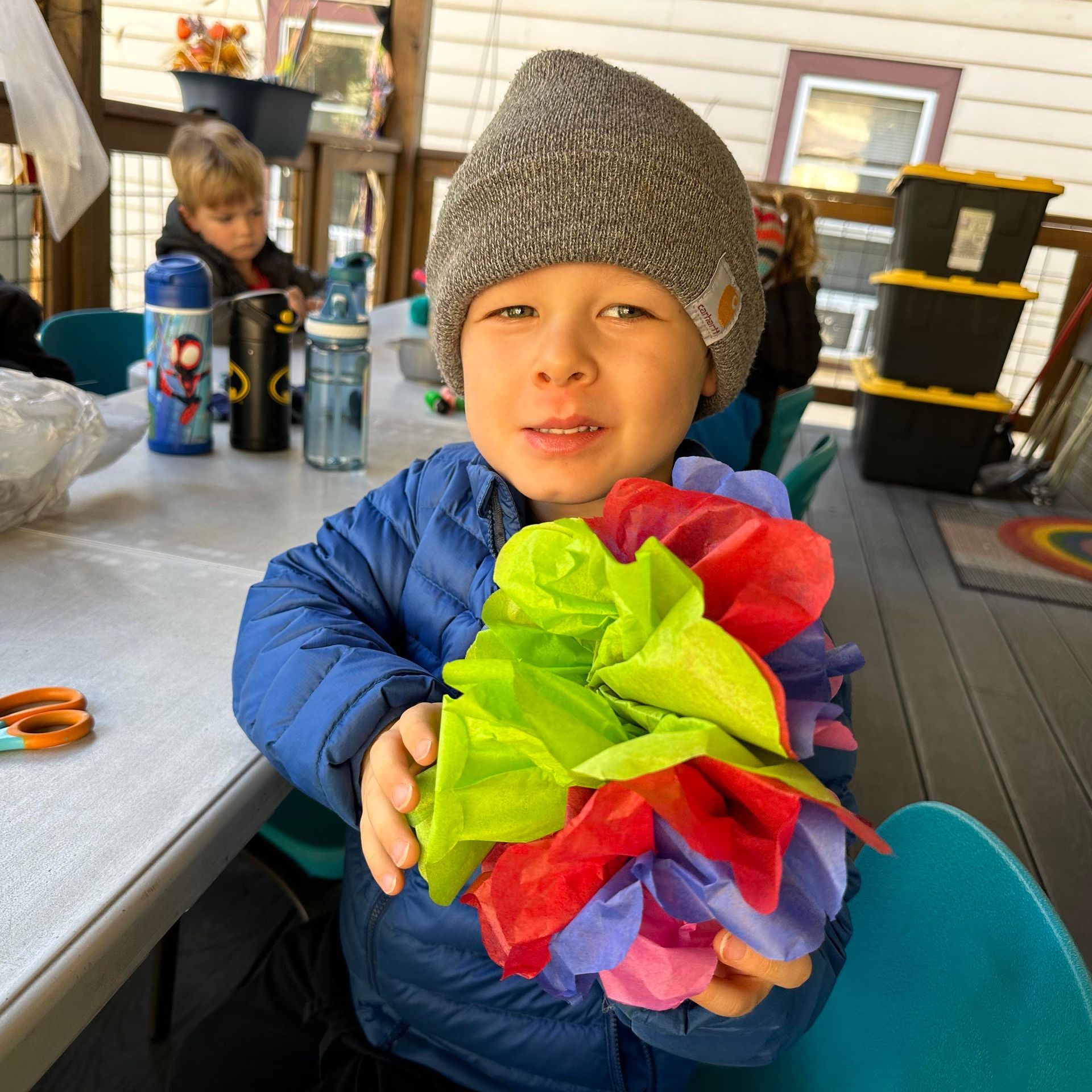 A child dressed in a jacket and hat sits ata table on an outdoor porch holding a bouquet of tissue paper flowers.