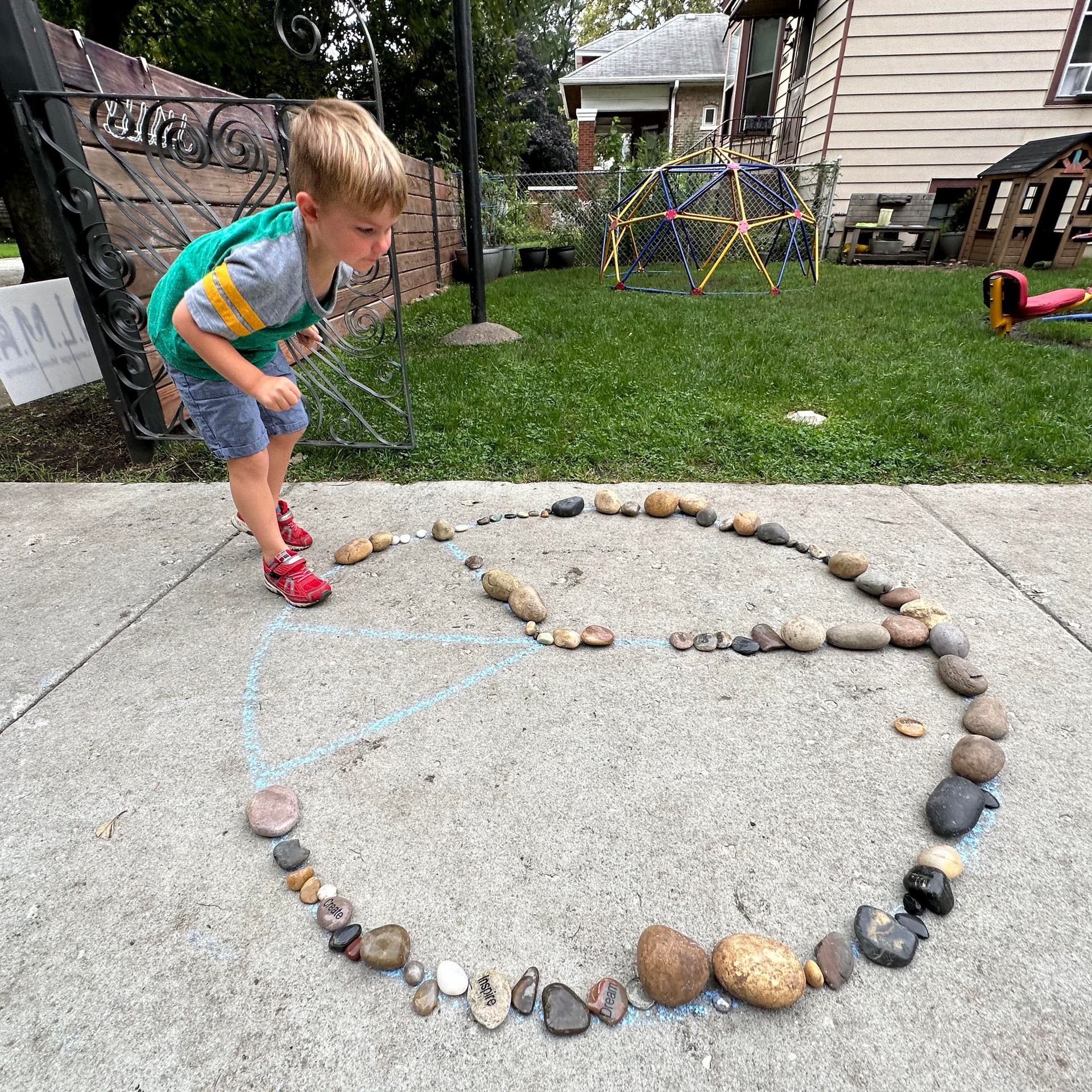 A child is outdoors creating a peace sign with rocks on the sidwalk.