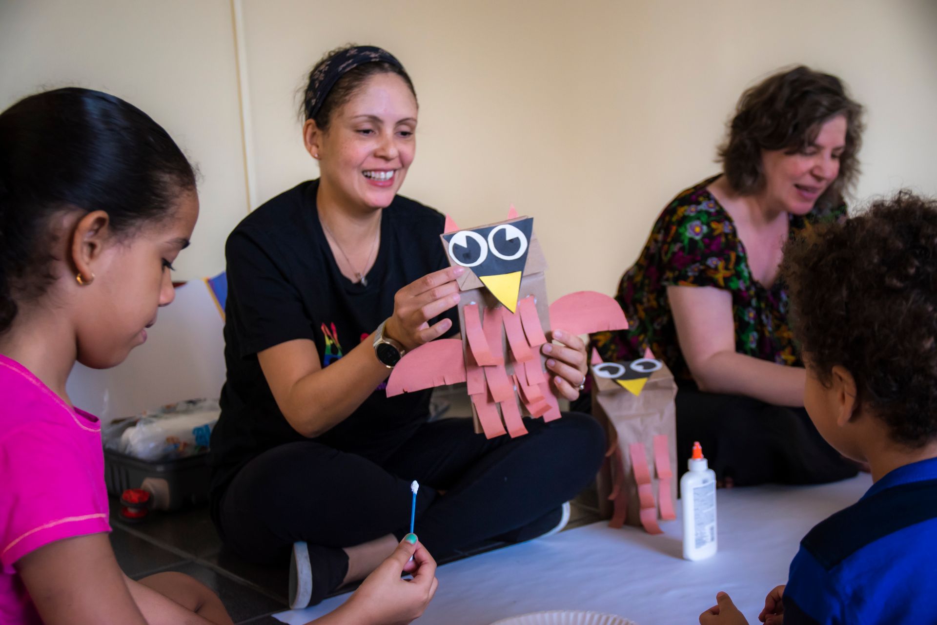 Two teachers seated on the floor with a small group of children, working together on an art project, which is making owls out of paper bags.