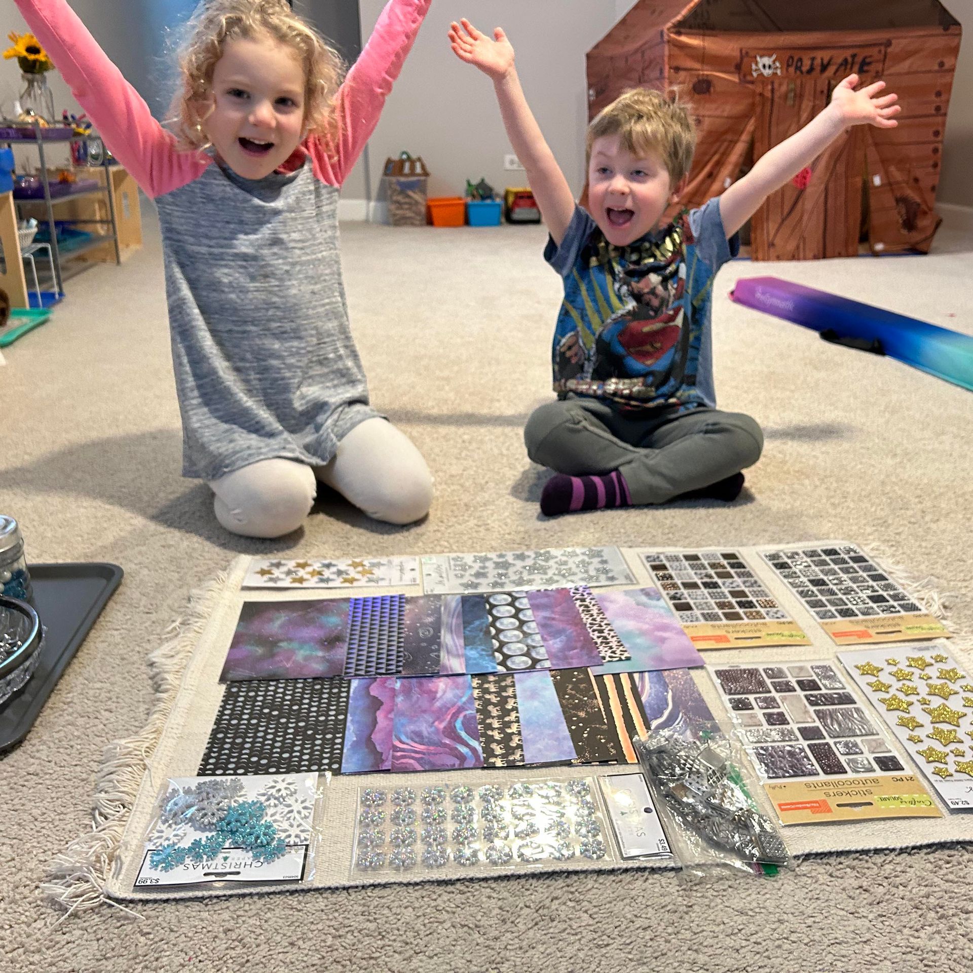Two children are seated on the floor in front of several paintings. They each have their arms outstretched over their heads in exaltation.