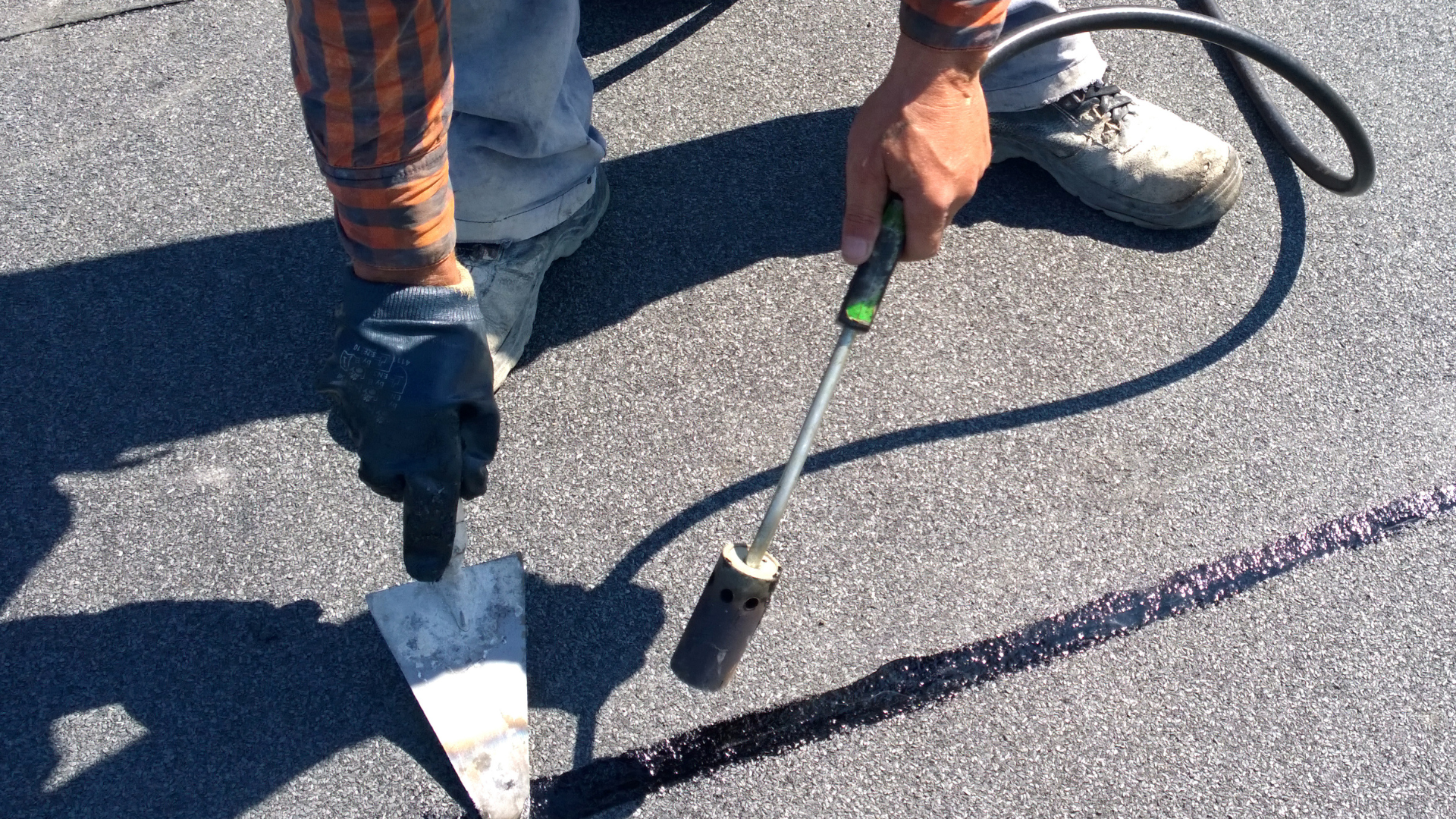 Person using a torch to seal a black asphalt roof with a trowel, outdoors.