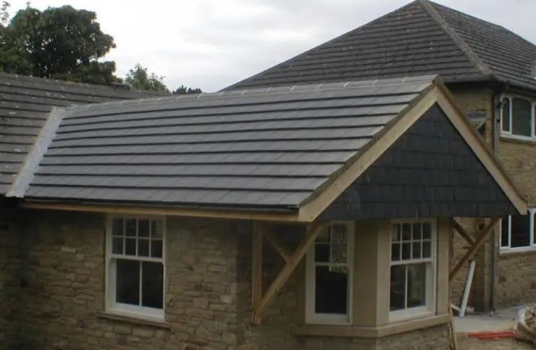 A house with a gray tiled roof extension, supported by wooden beams, over windows on a stone facade.