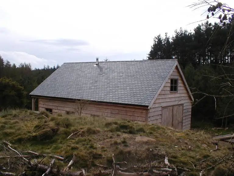 Wooden shed with gray roof, set in a grassy, wooded area under an overcast sky.