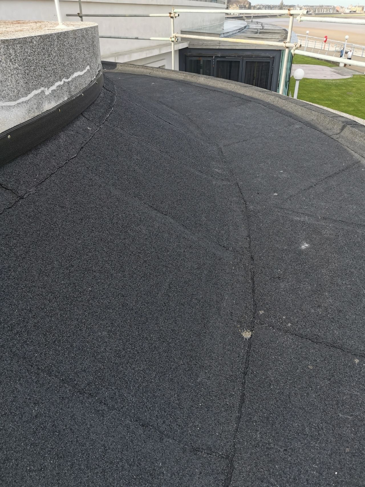 Curved, dark asphalt roof with cracks, viewed from a rooftop, with a building and sky in the background.