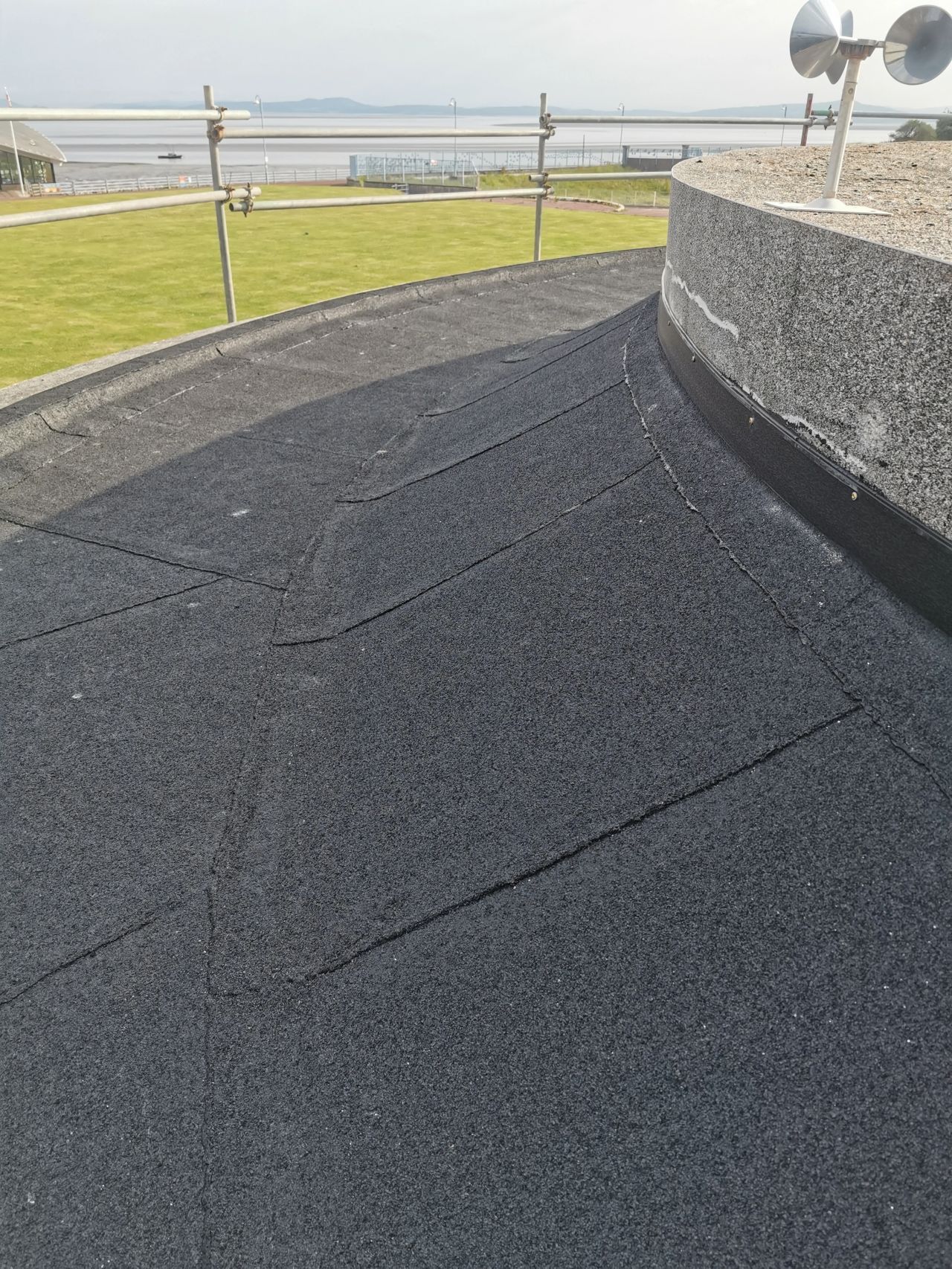 Black rubber roof with lines, curving around a wall, with a grassy area and water in the background.