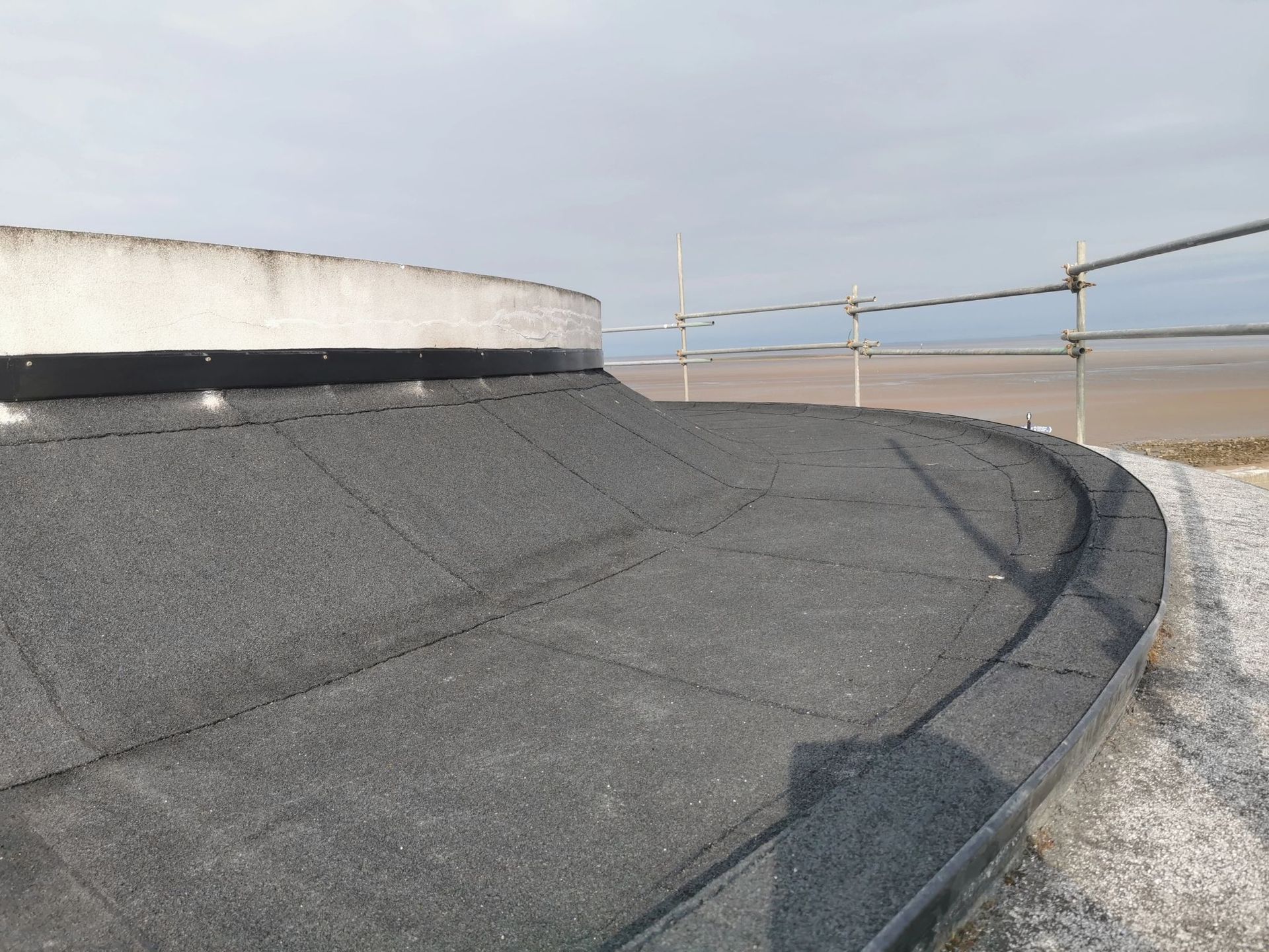 A flat, curved roof covered in dark gray material. Scaffolding and the ocean are visible in the background.