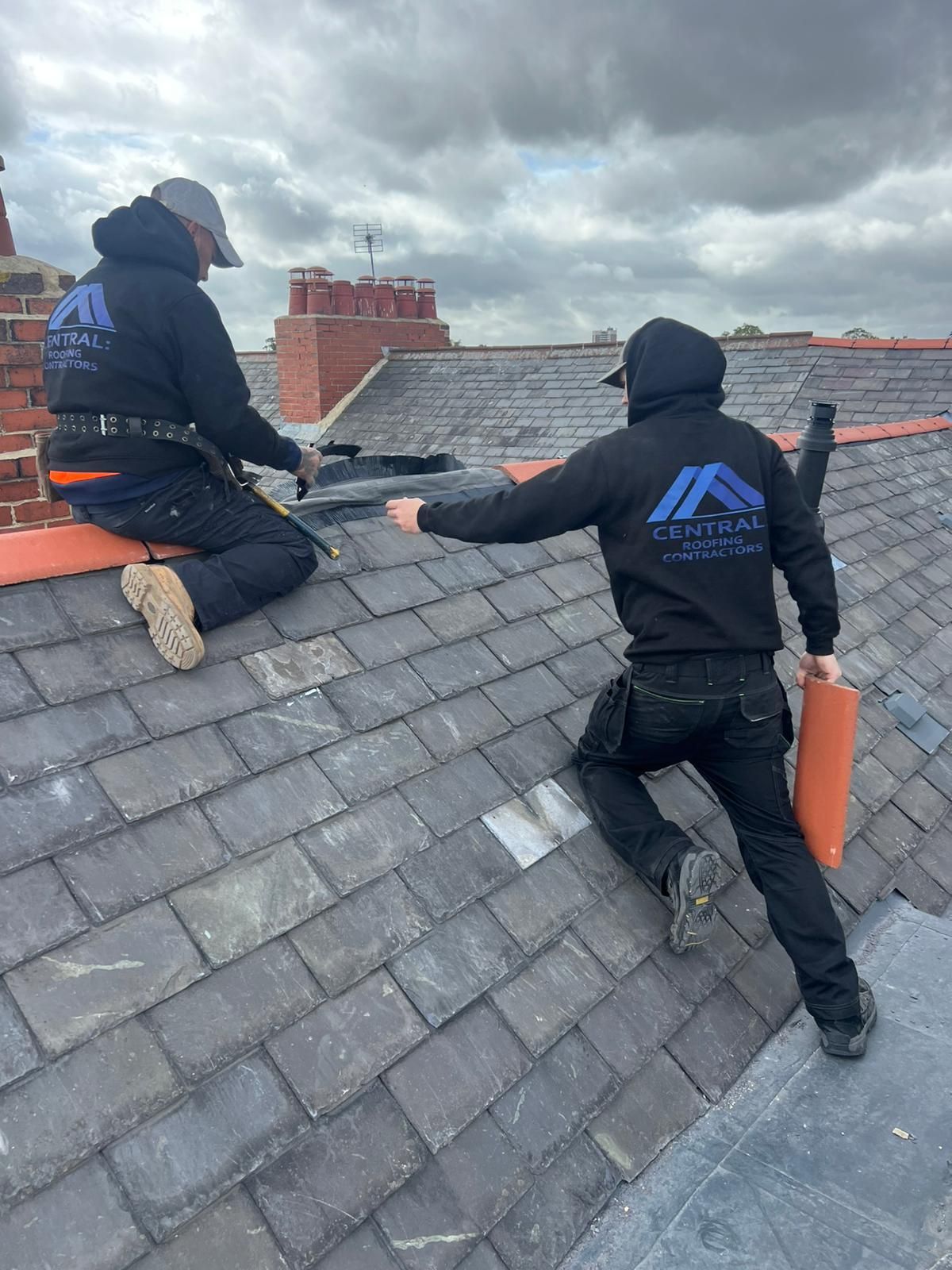 Two roofers on a slate roof, one holding a roll, overcast sky.