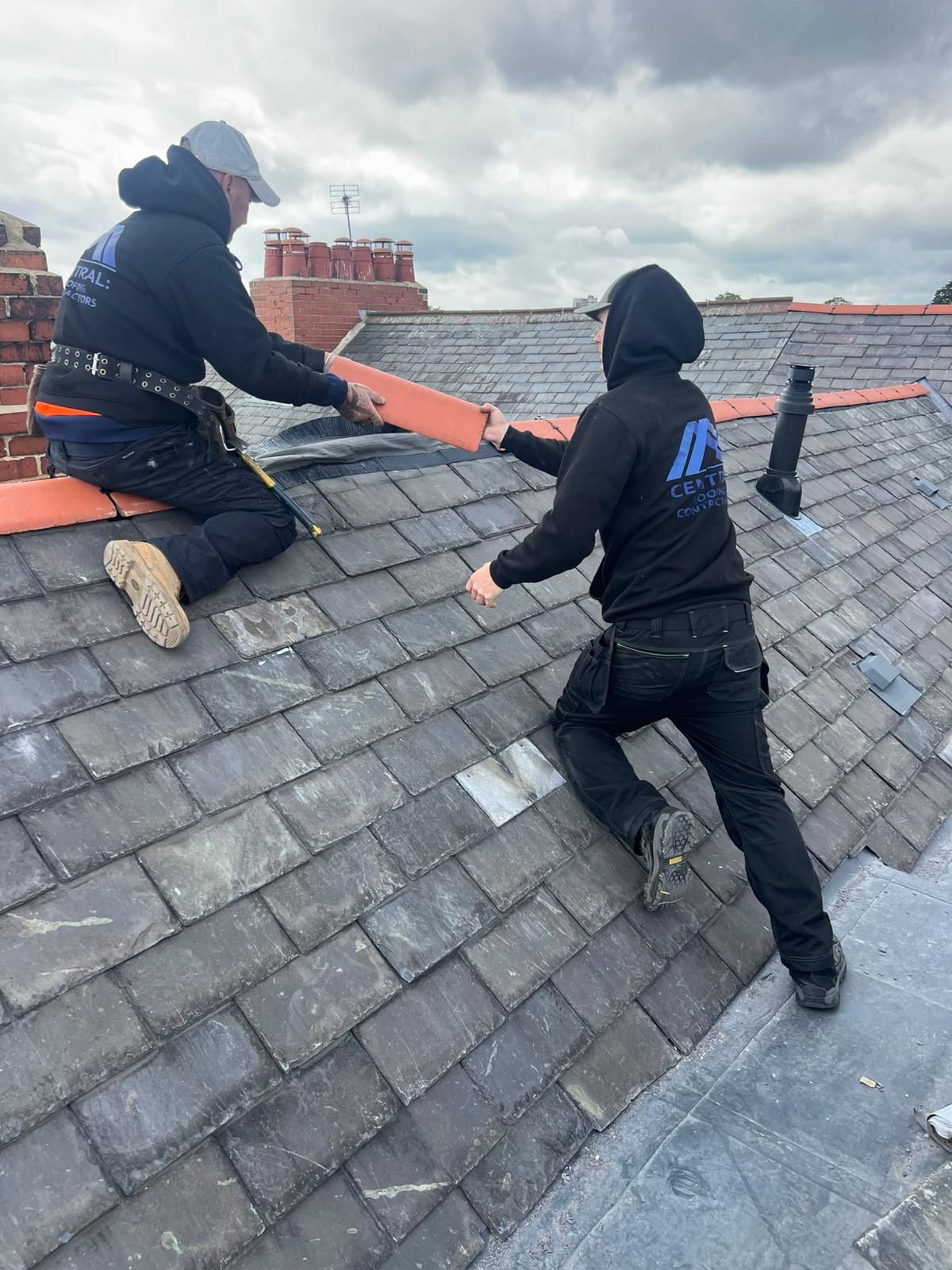 Two roofers, in black hoodies, on a slate roof, replacing a section near a chimney.