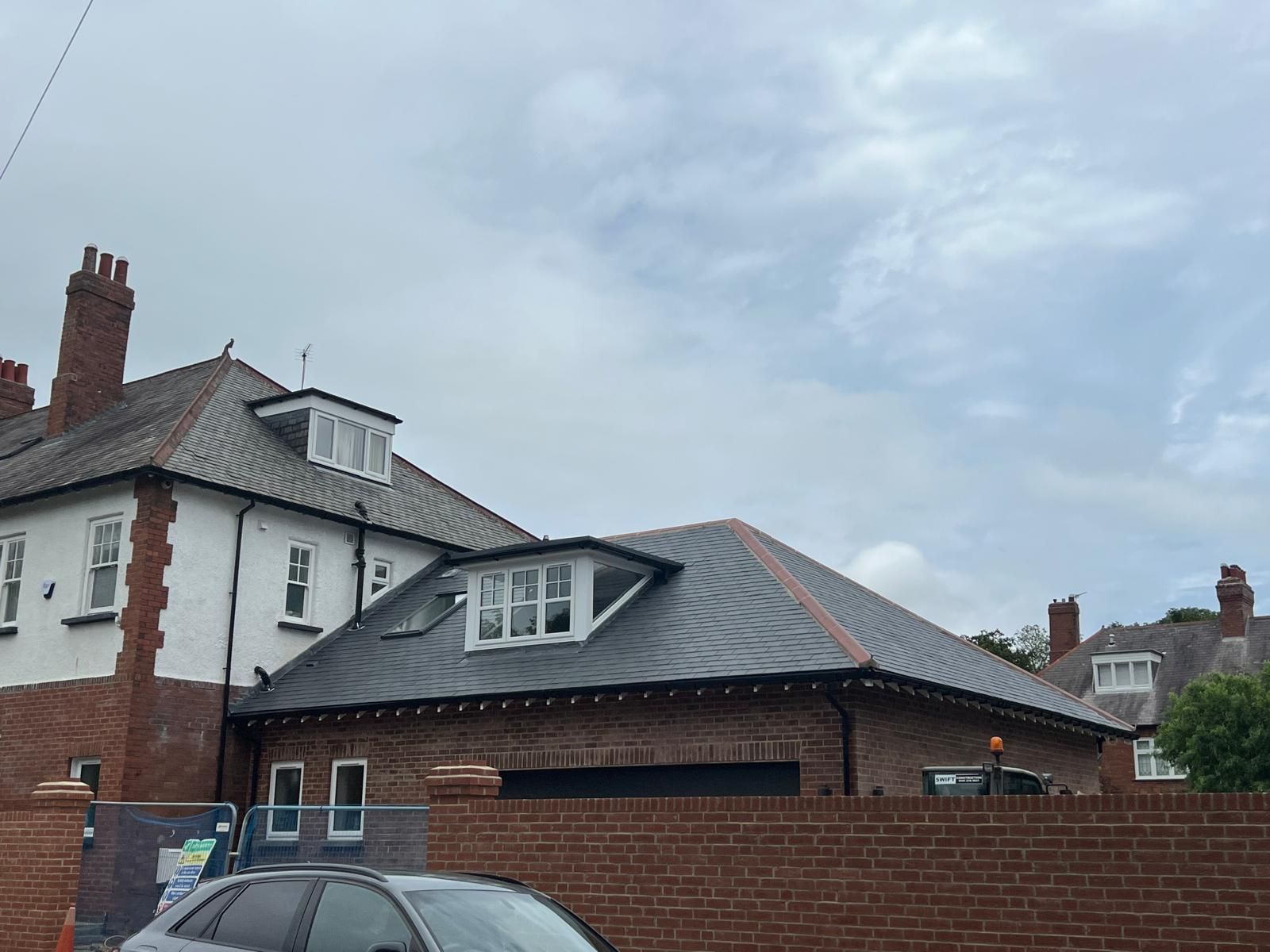 Brick building with a slate roof and dormer windows, behind a brick wall under a cloudy sky.