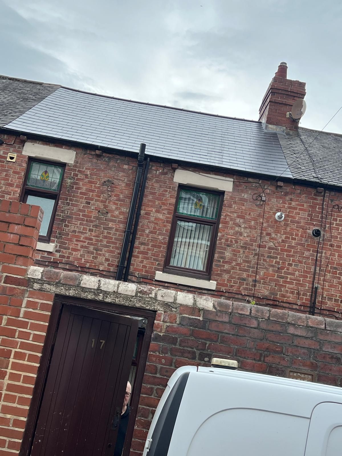 Brick house with two windows, a chimney, and a black pipe. A door and a white van are visible.